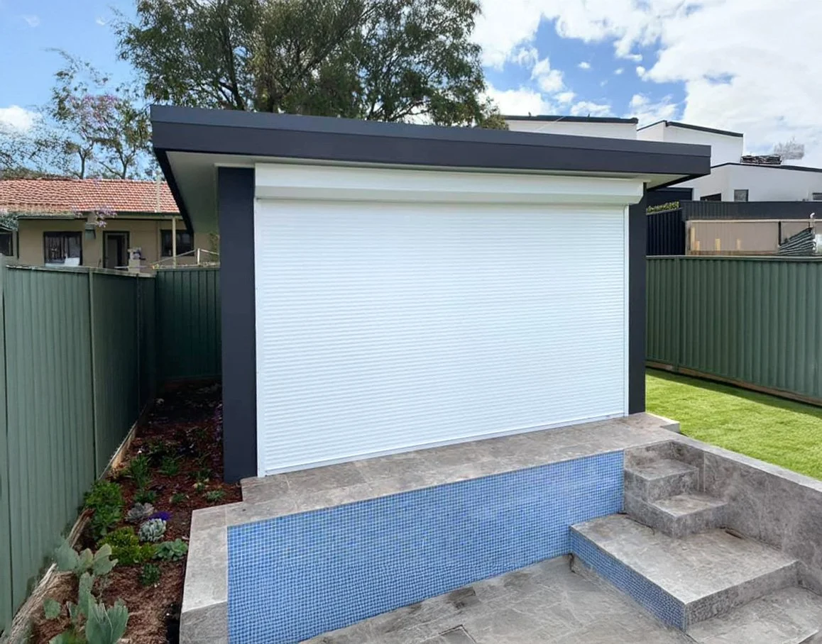 A backyard with a small modern structure featuring a white roll-up door, a tiled pool with blue mosaic tiles, stone steps, and a fenced yard with grass and plants under a partly cloudy sky.