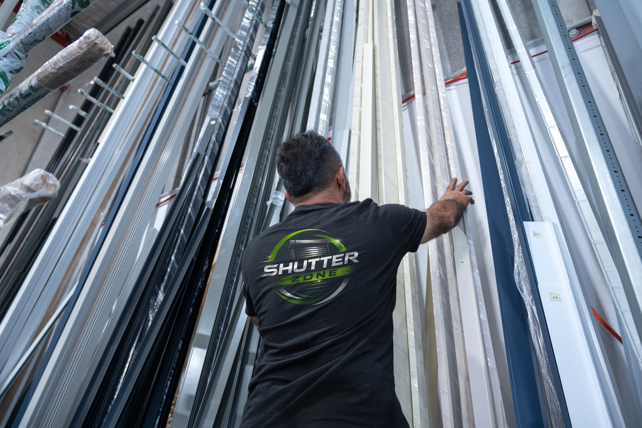 A man wearing a black T-shirt with the words 'Shutter Zone' on the back is selecting or inspecting window frames or shutters on a wall of various window parts in a warehouse in Sydney, NSW.