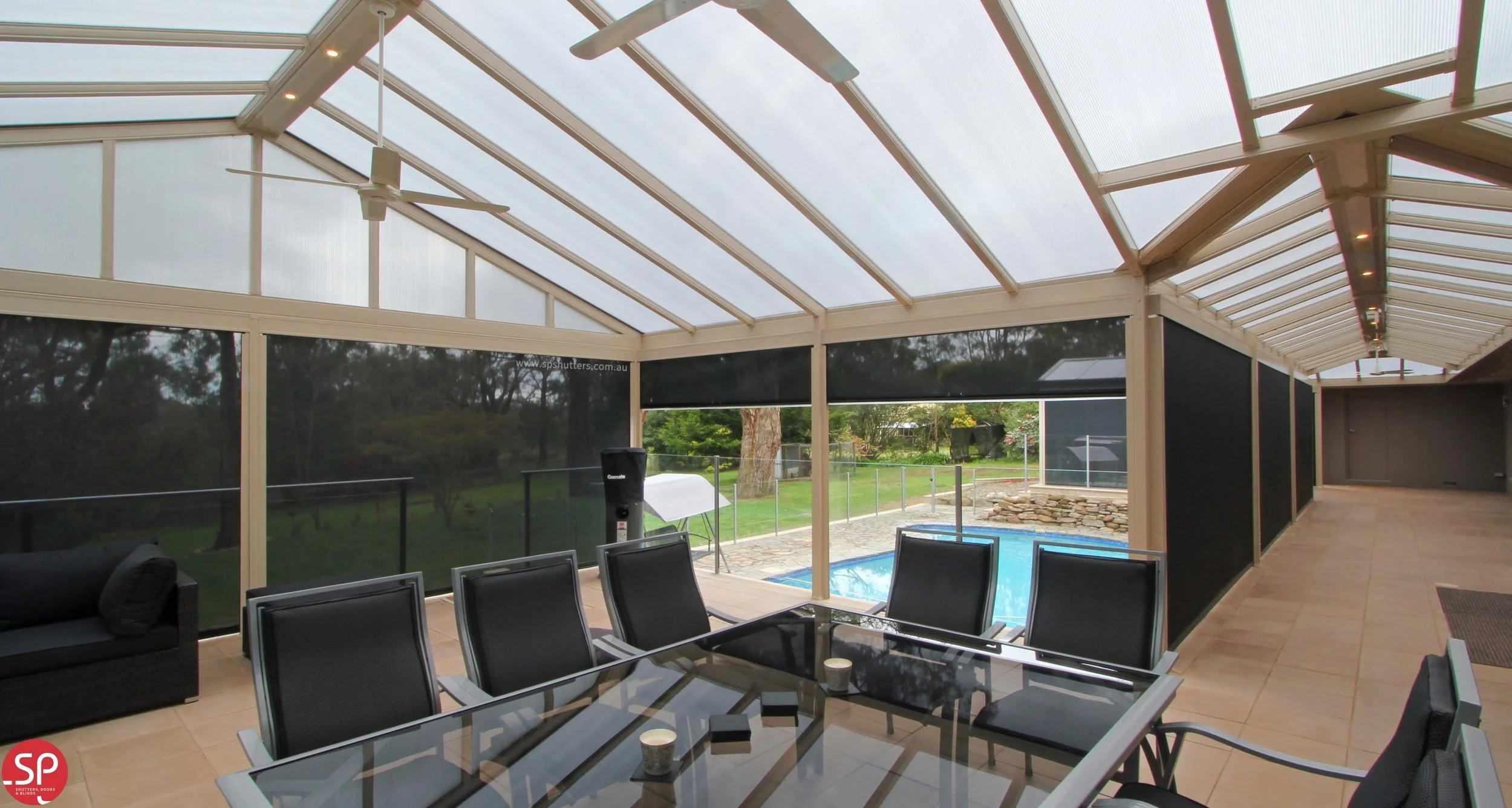 Enclosed patio by eZip shutters with ceiling fans, glass-topped dining table with six chairs, black sofa, and a view of a backyard with a pool and green yard. Located in Sydney, Australia.