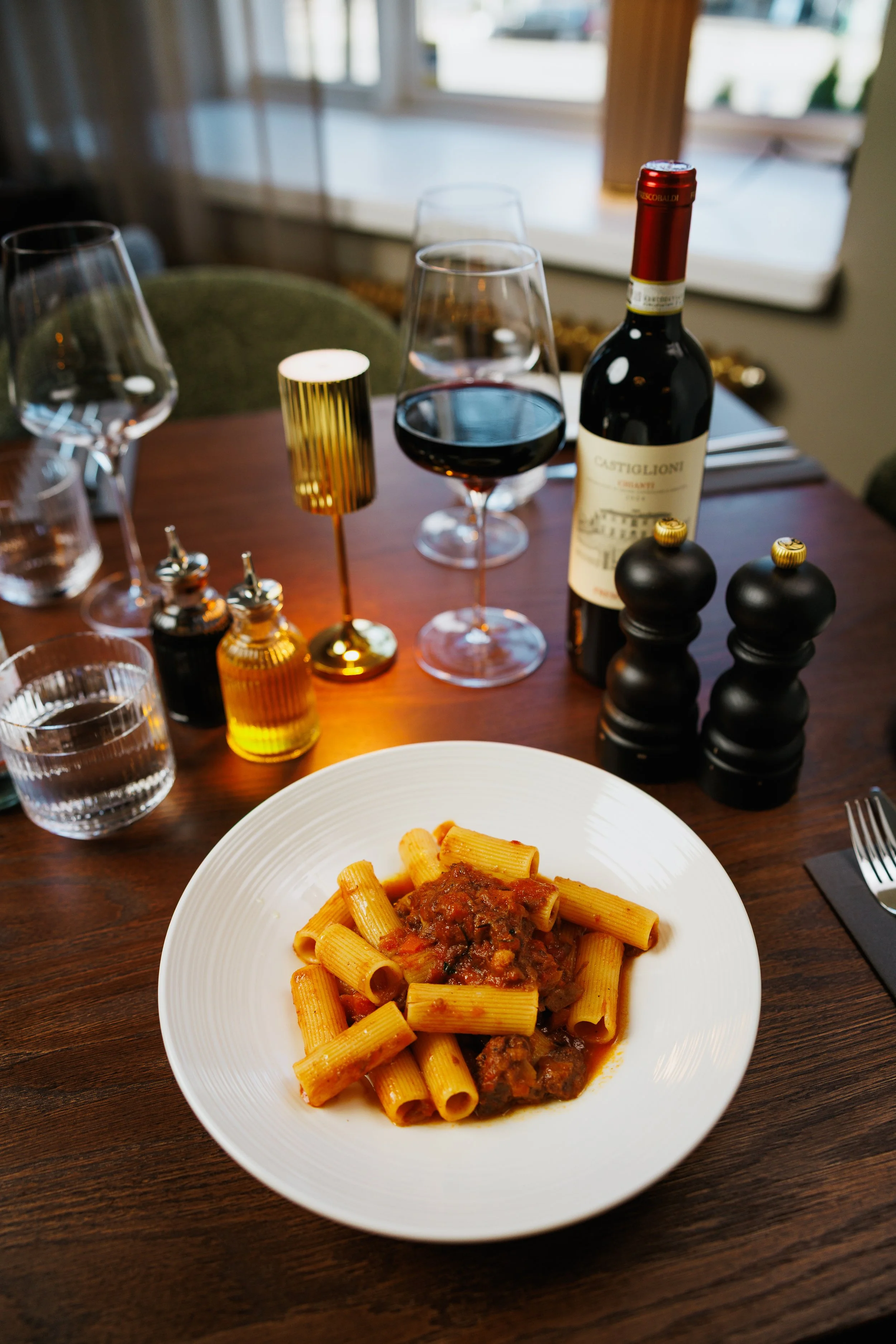 A plate of rigatoni pasta with meat sauce on a wooden dining table, with a glass of red wine, a bottle of red wine, water glasses, salt and pepper shakers, and decorative candles.