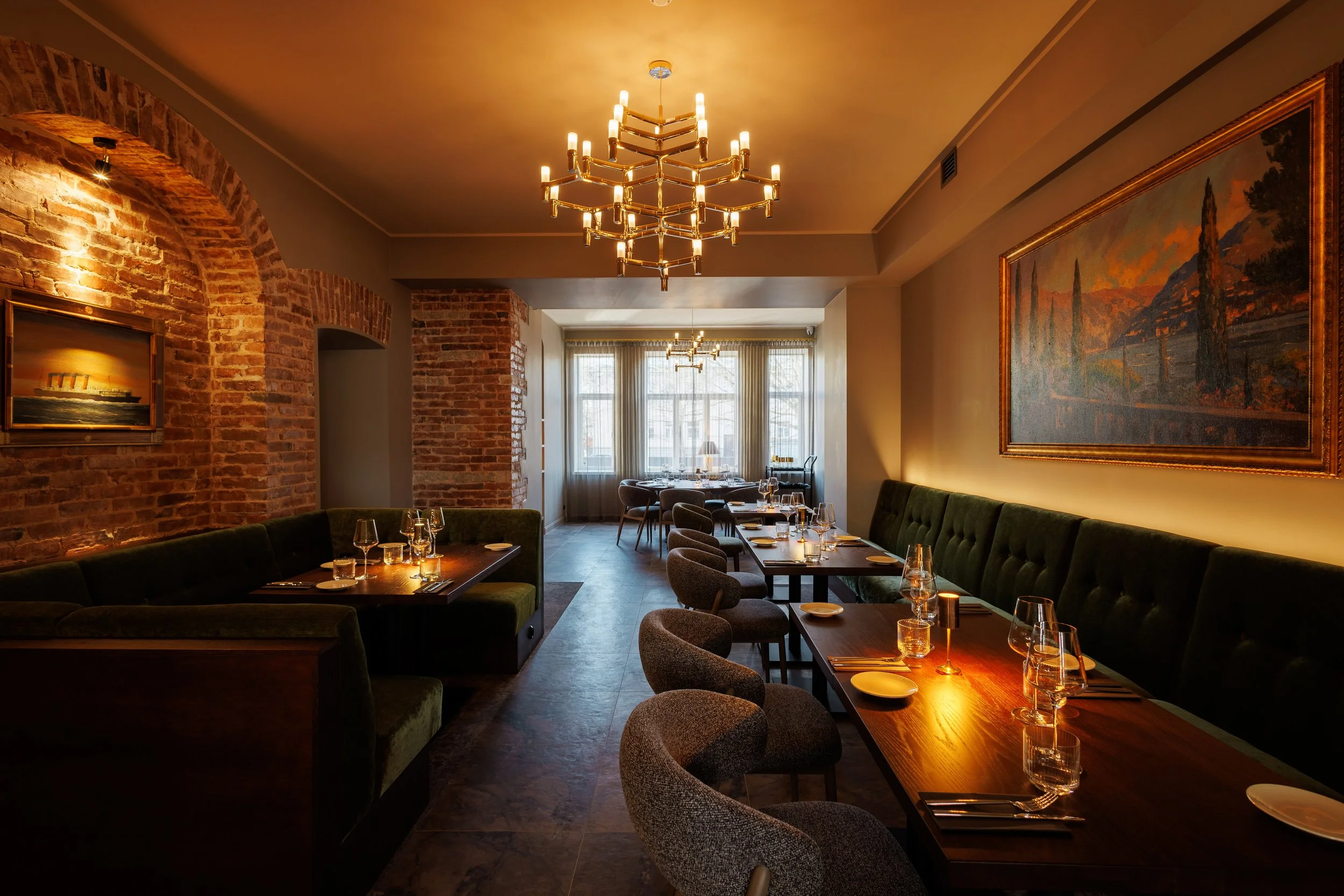 A warmly lit restaurant dining area with exposed brick walls, modern chandeliers, and tables set for a meal with glassware and napkins.