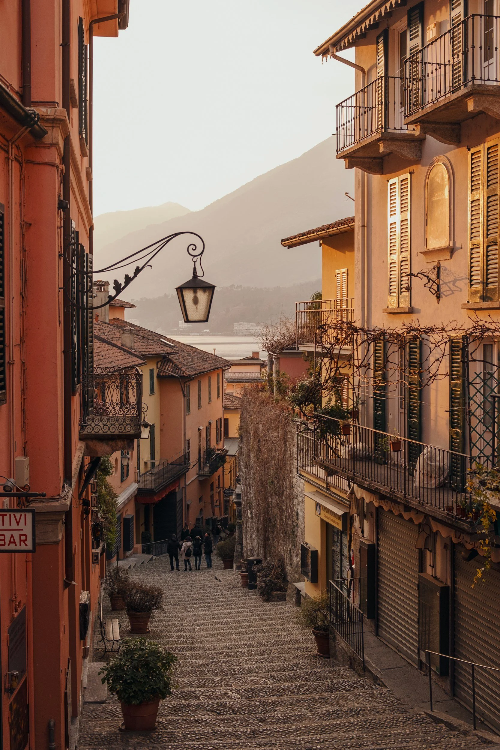 A narrow cobblestone street in a European town at sunset, with colorful buildings, iron balconies, potted plants, and a few pedestrians walking downhill, mountains in the background.