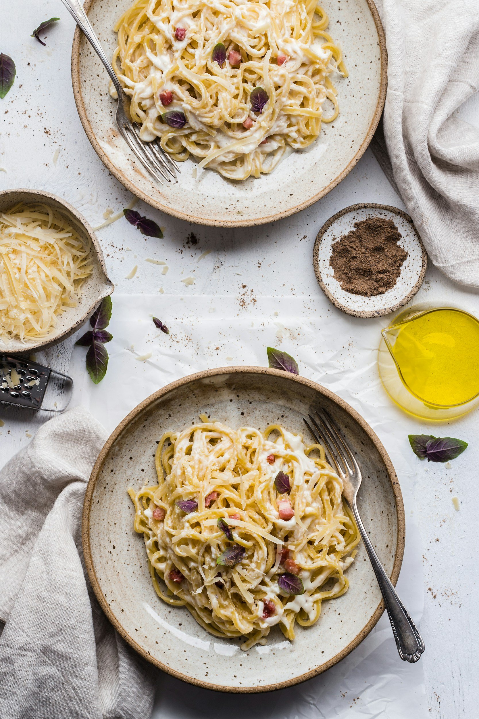 Two plates of creamy pasta with bacon bits and purple basil leaves, surrounded by grated cheese, ground black pepper in a small dish, a pitcher of olive oil, and a cheese grater on a white table with a beige linen napkin.