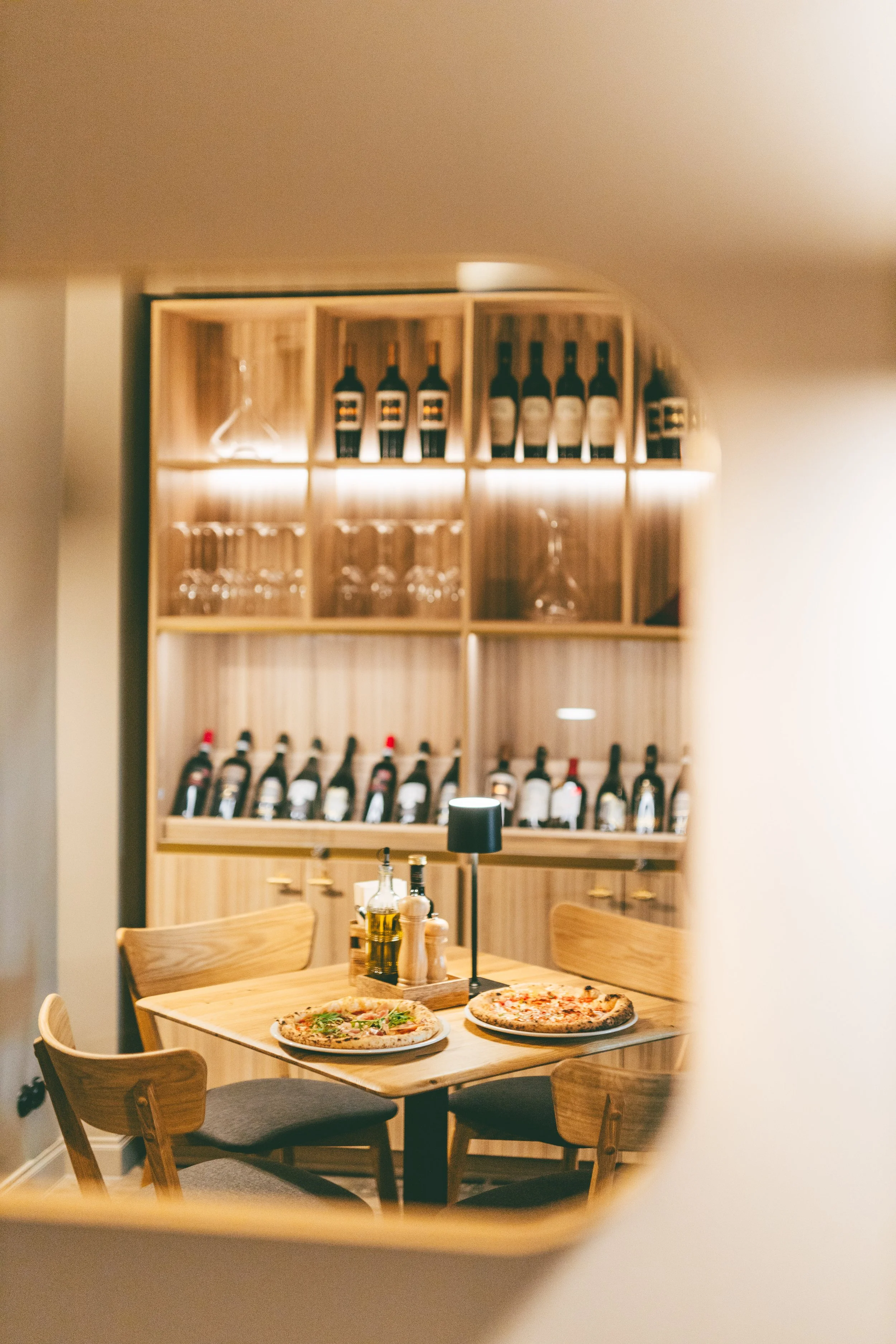 Two pizzas on a wooden table with condiments, surrounded by four chairs in a cozy restaurant, viewed through a round opening in the wall, with a wooden wine rack and glasses in the background.