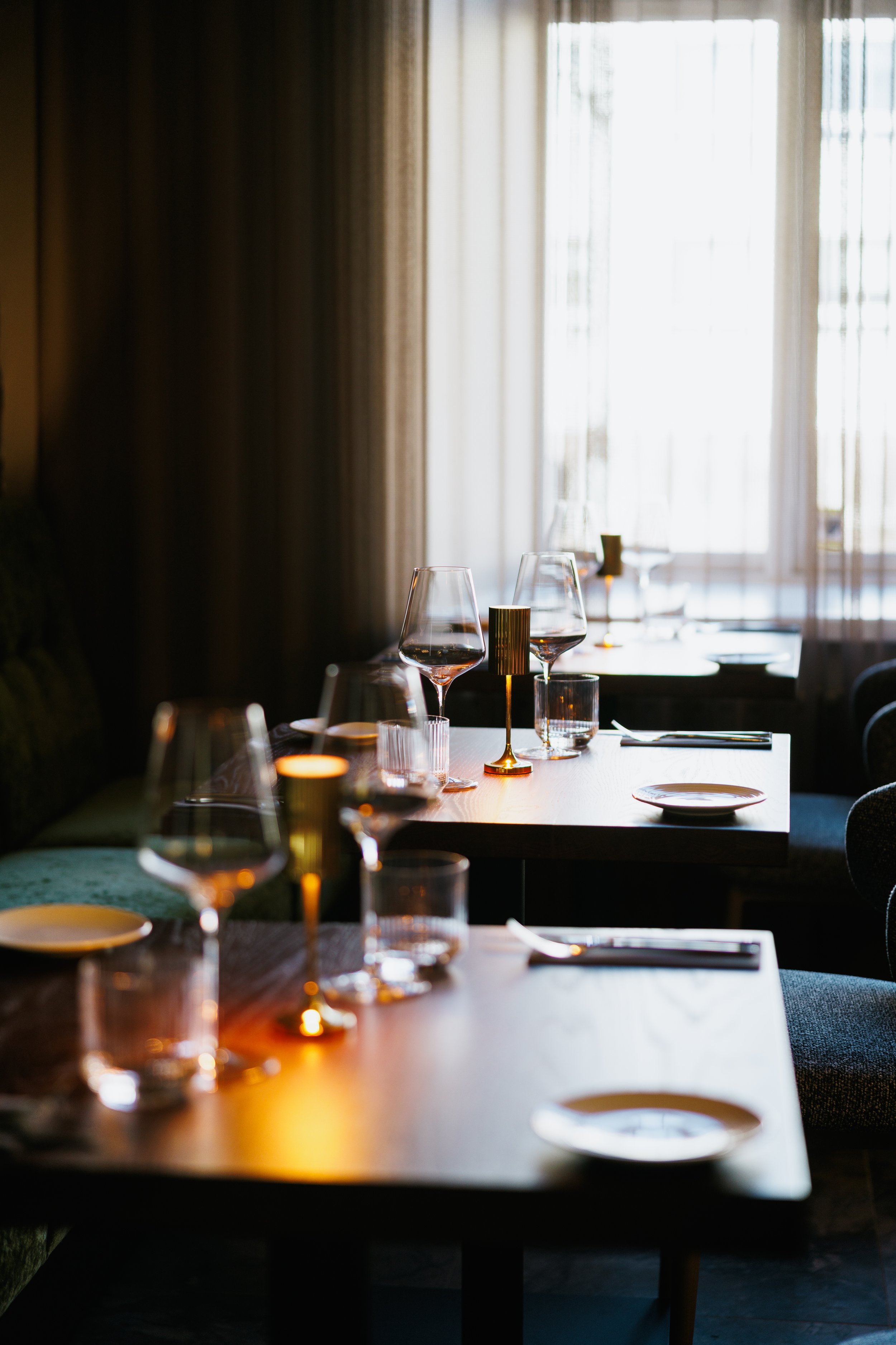 Elegant restaurant table setup with wine glasses, candles, plates, and cutlery near a window with natural light.