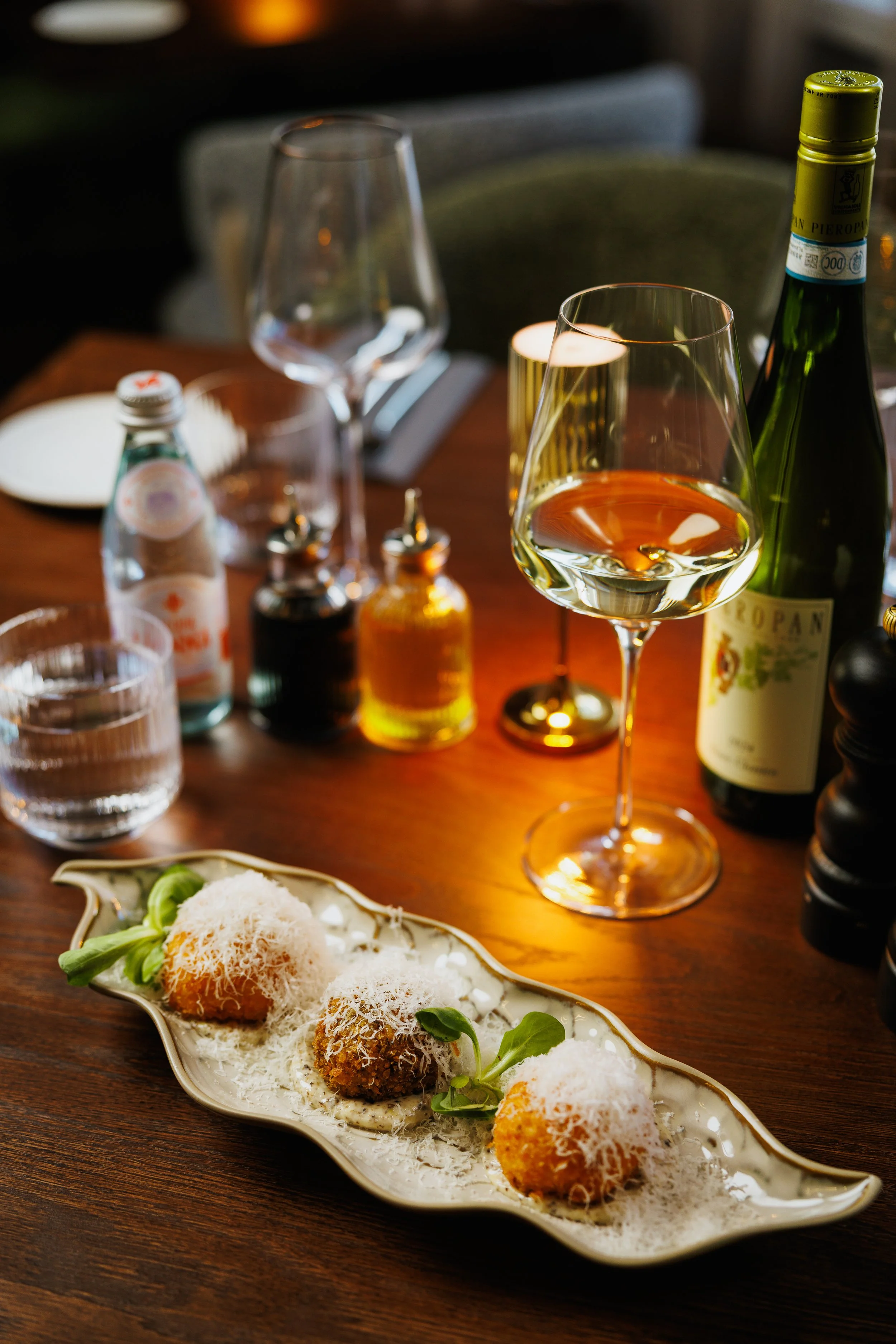 Elegant table setting with three arancini balls topped with grated cheese and microgreens on a decorative white plate, surrounded by wine glasses, a bottle of white wine, water glasses, and condiment bottles, in a softly lit restaurant.