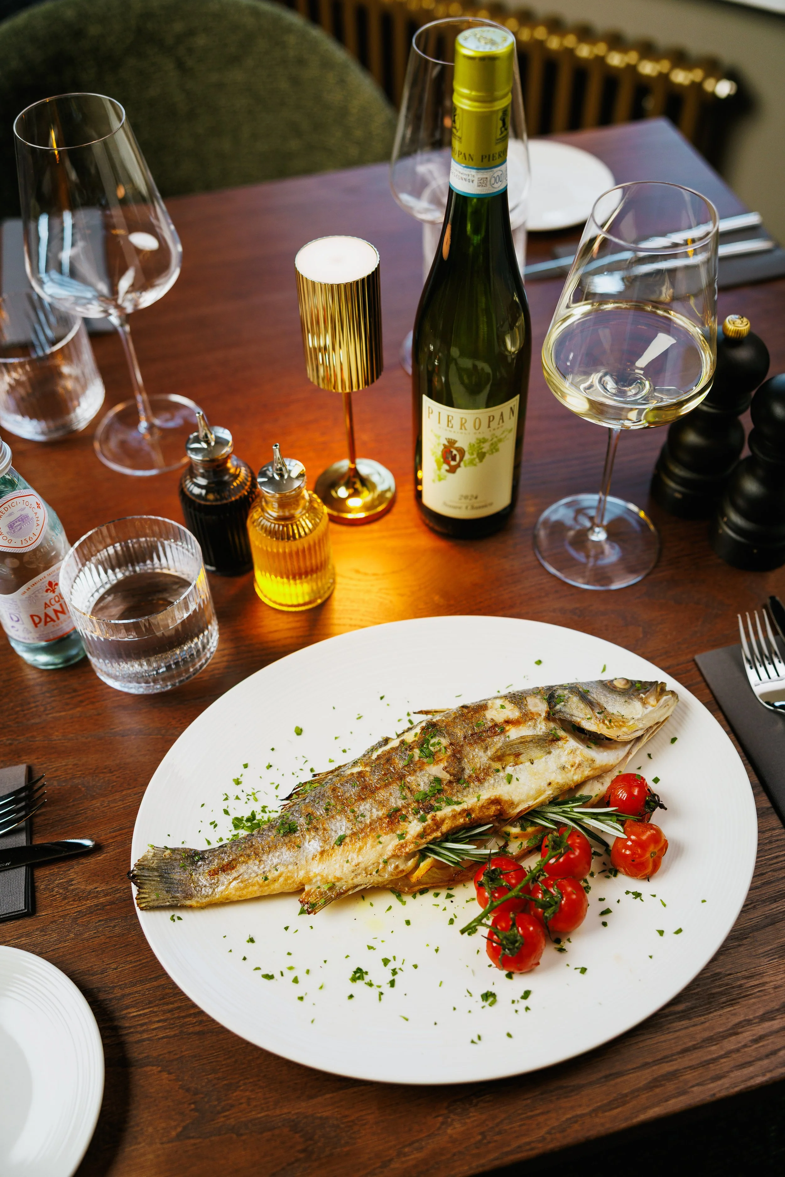 A table set with a roasted whole fish garnished with herbs and cherry tomatoes, alongside a glass of white wine, a bottle of Pieropan wine, empty wine glasses, water glasses, a candle, and seasoning shakers.