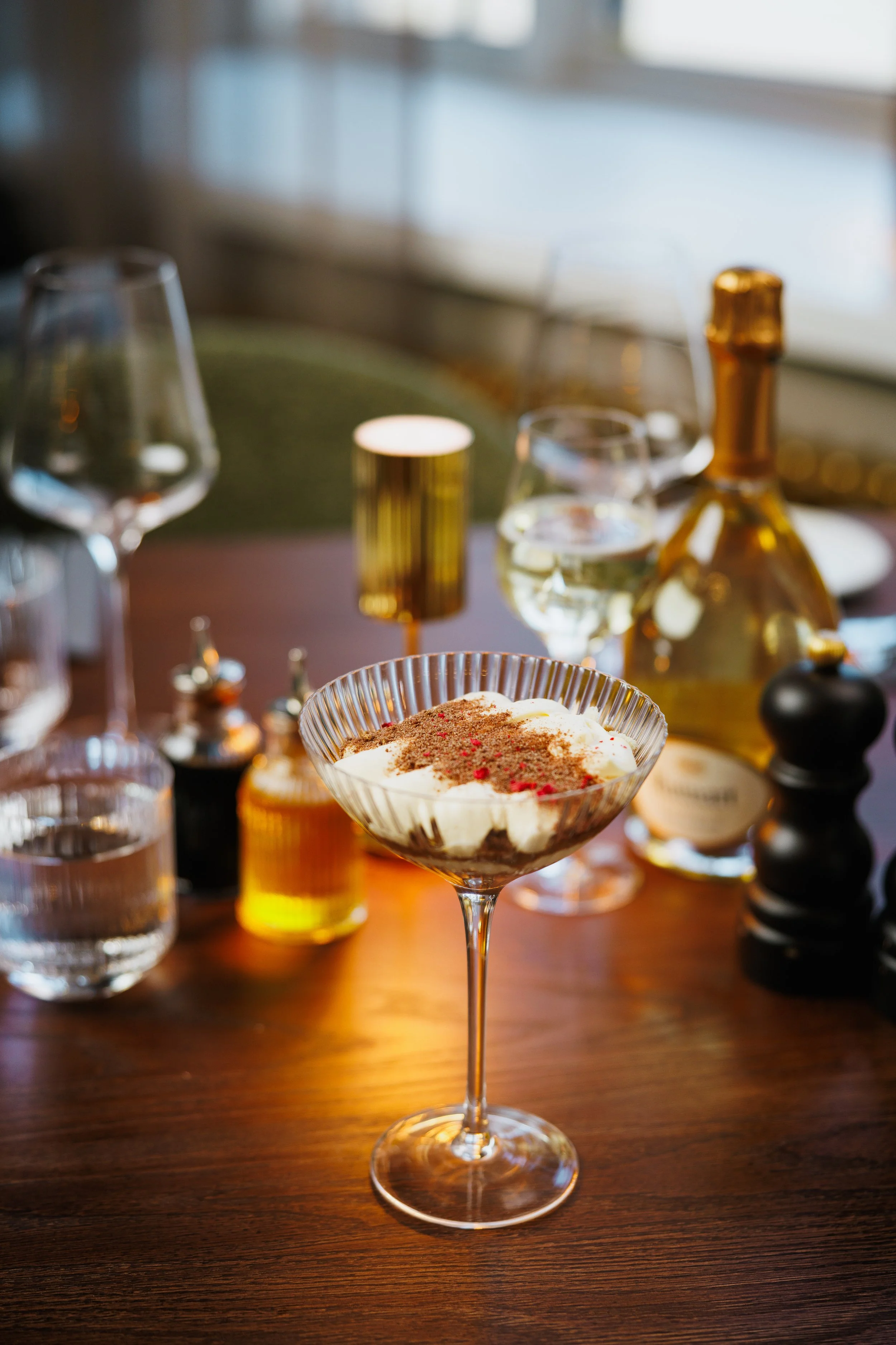 Dessert in a cocktail glass on a wooden table with liquor bottles, glasses of water, and candleholders in the background.