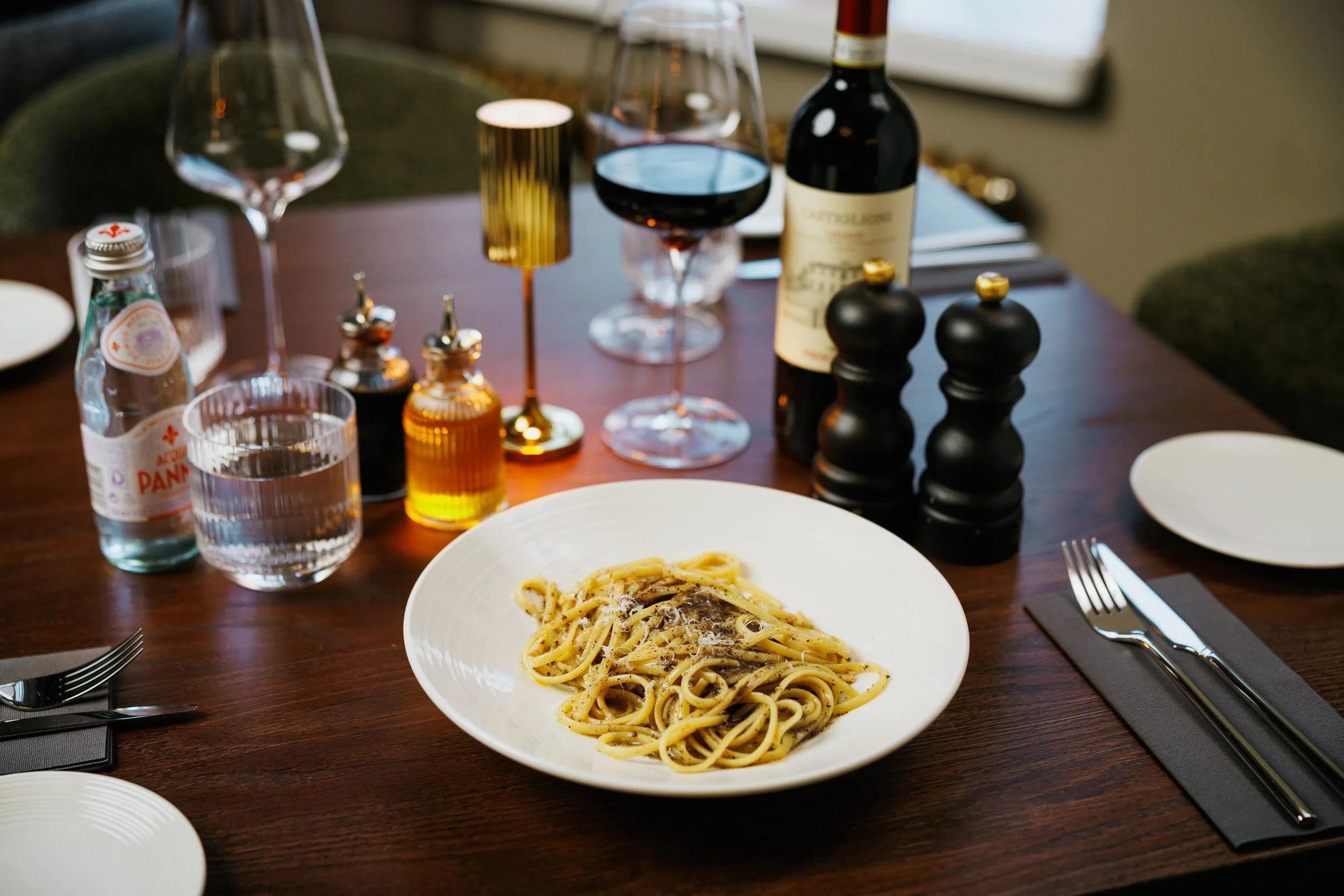 A dining table set for a meal featuring a plate of spaghetti with sauce, a glass of red wine, a bottle of red wine, a water bottle, a glass of water, various condiments, salt and pepper shakers, and wine glasses, with black utensils on a napkin.