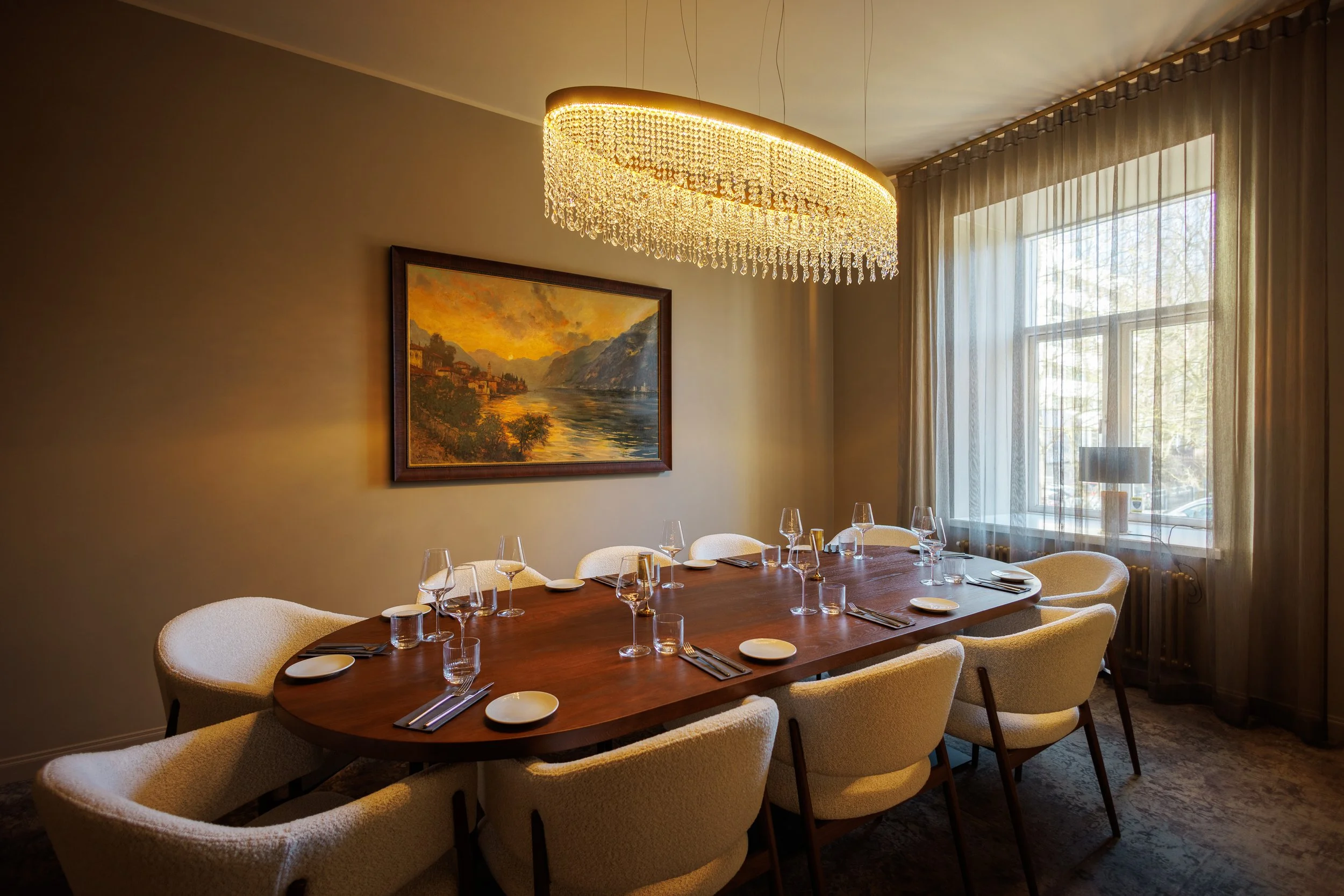 A dining room with a large wooden table set for dinner, surrounded by cream-colored upholstered chairs, under a sparkling chandelier, with a landscape painting on the wall, and a window with sheer curtains allowing natural light in.