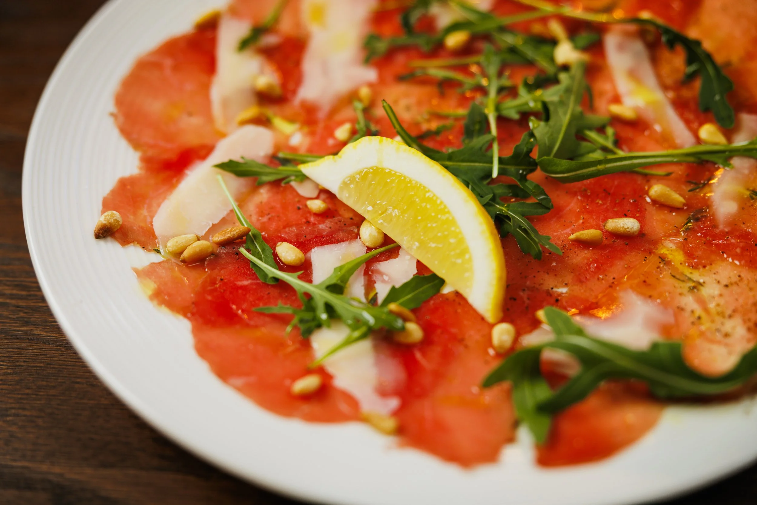 A plate of thinly sliced smoked salmon garnished with arugula, pine nuts, lemon wedge, and shavings of Parmesan cheese.