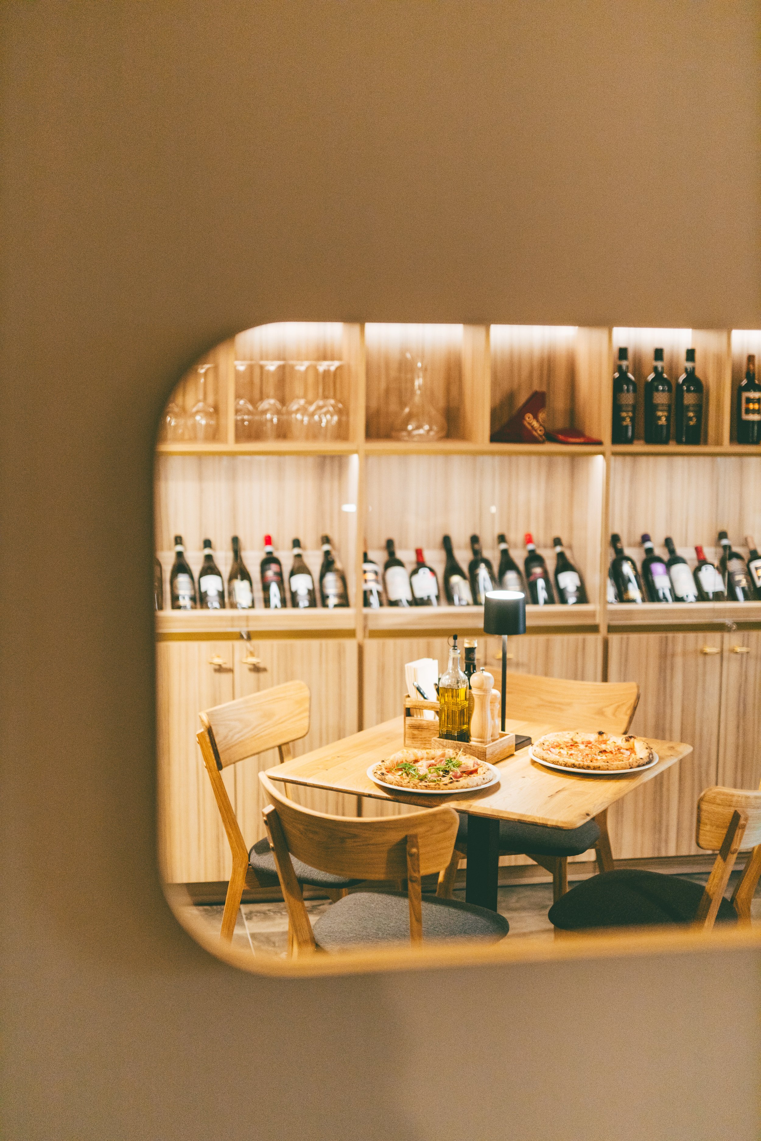 A cozy restaurant interior with wooden table and chairs, two pizzas, and a wine shelf in the background.