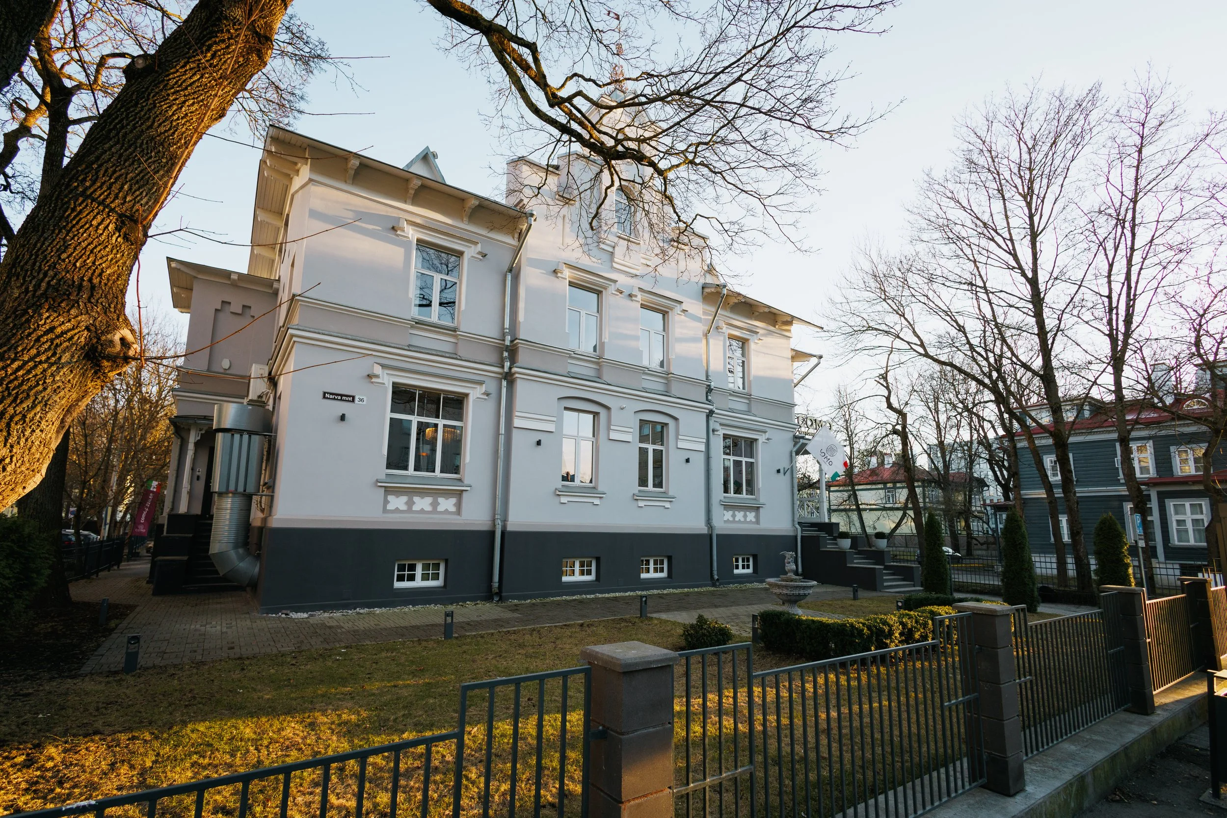 A white multi-story house with decorative window frames and gray base, surrounded by leafless trees, a black metal fence, and a small garden with a fountain, in a neighborhood during late afternoon or early evening.