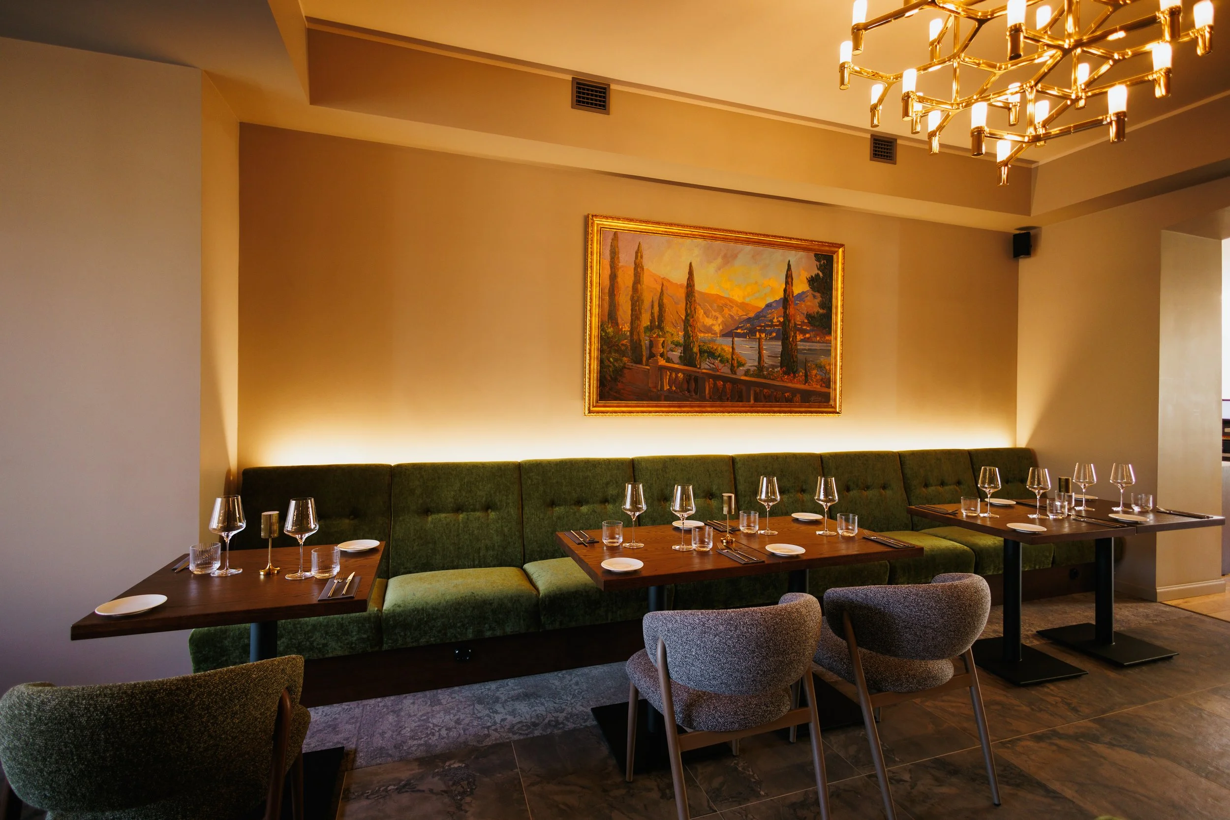 Empty restaurant table set with wine glasses, water glasses, plates, and utensils in front of a green upholstered banquette and a framed landscape painting on the wall.