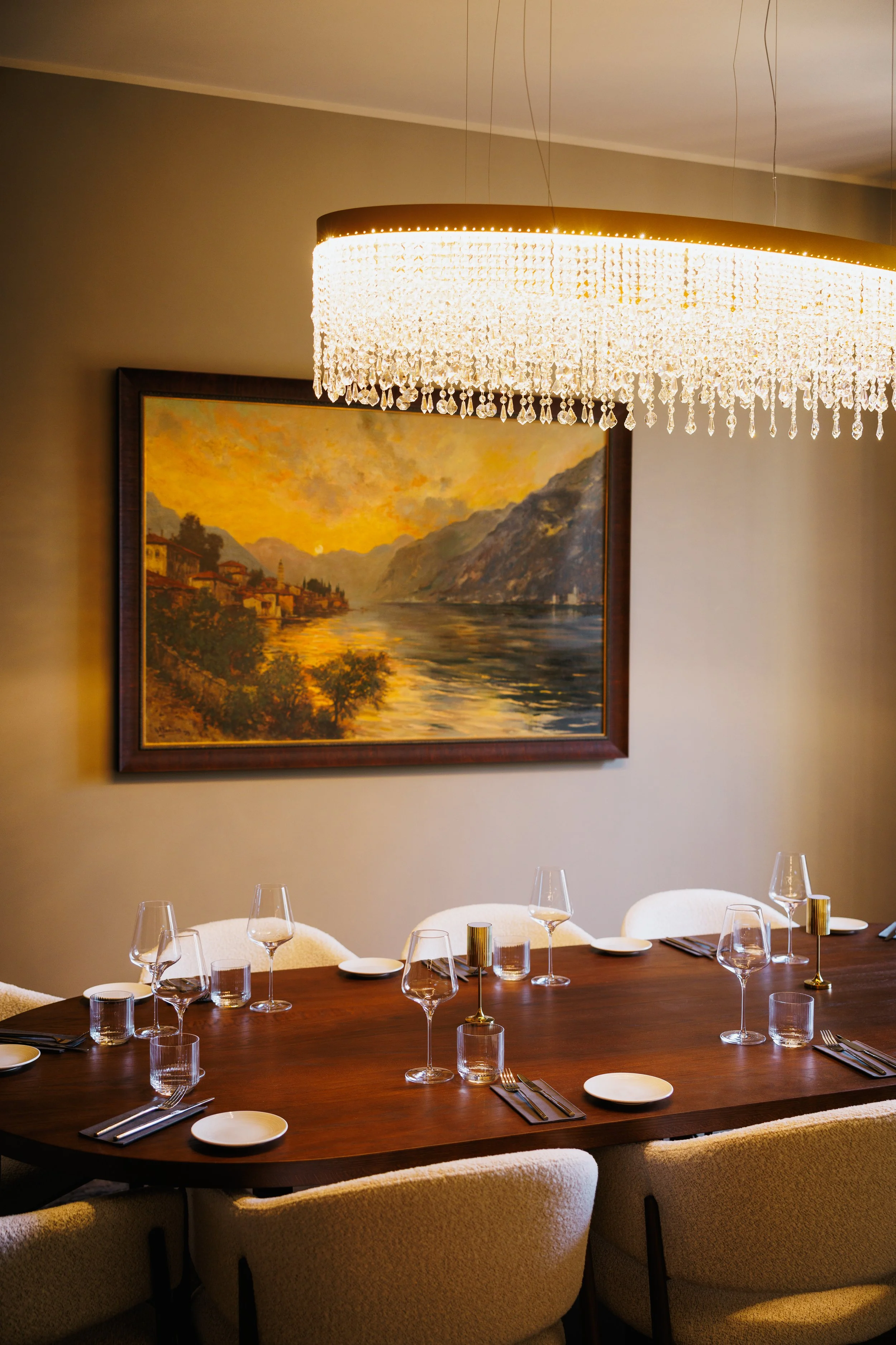 A dining room with a large, dark wood table set with glassware and plates, creamy upholstered chairs, a framed landscape painting, and a chandelier with hanging crystals.