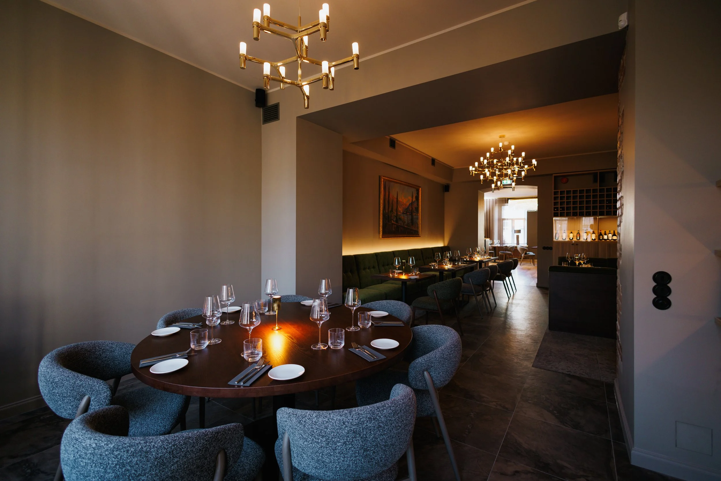 Interior view of a modern restaurant with dim lighting, featuring a round dining table set with glasses, plates, and silverware, surrounded by upholstered chairs, with additional tables, paintings, chandeliers, and a bar area in the background.
