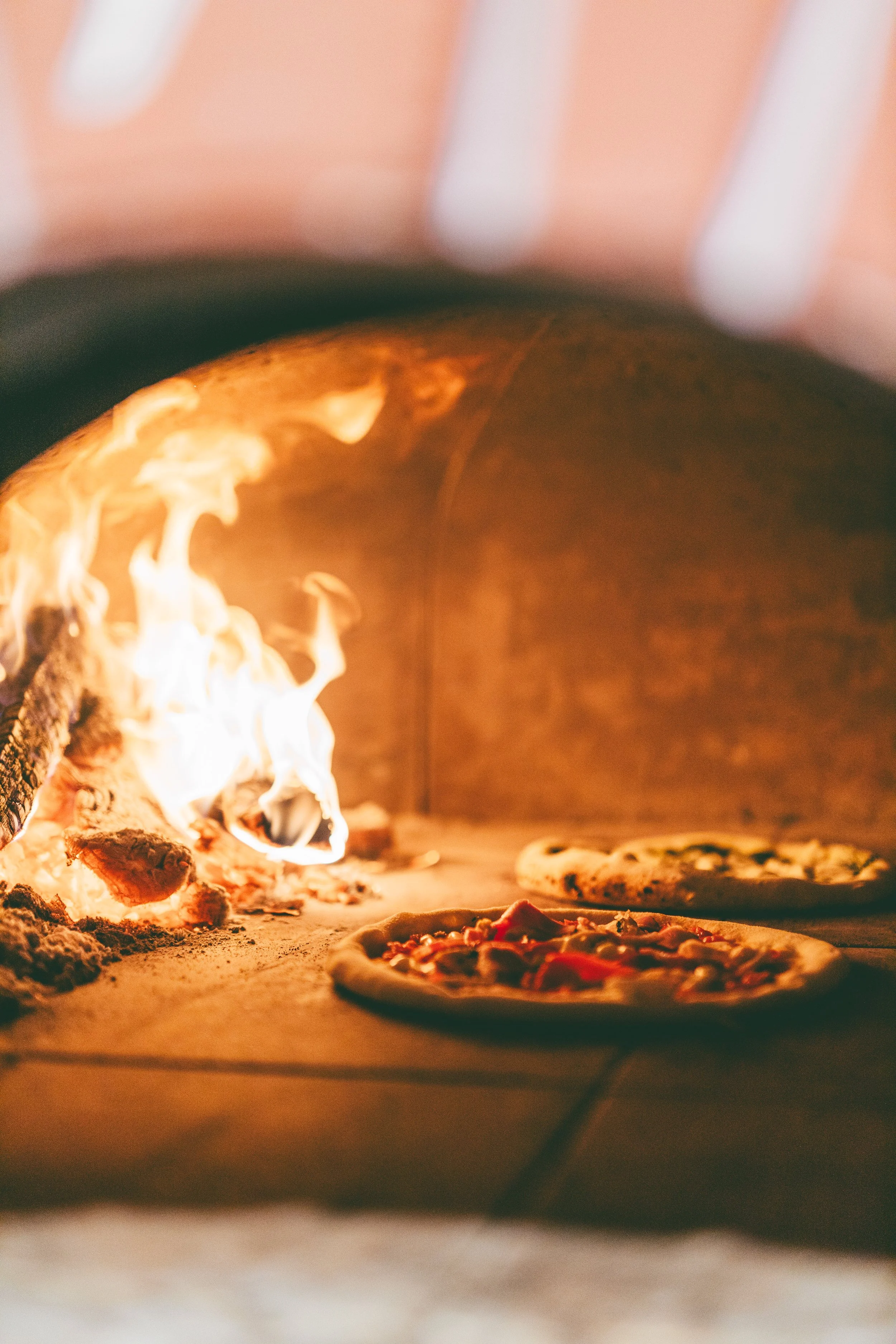 Two pizzas baking inside a wood-fired oven with flames visible.