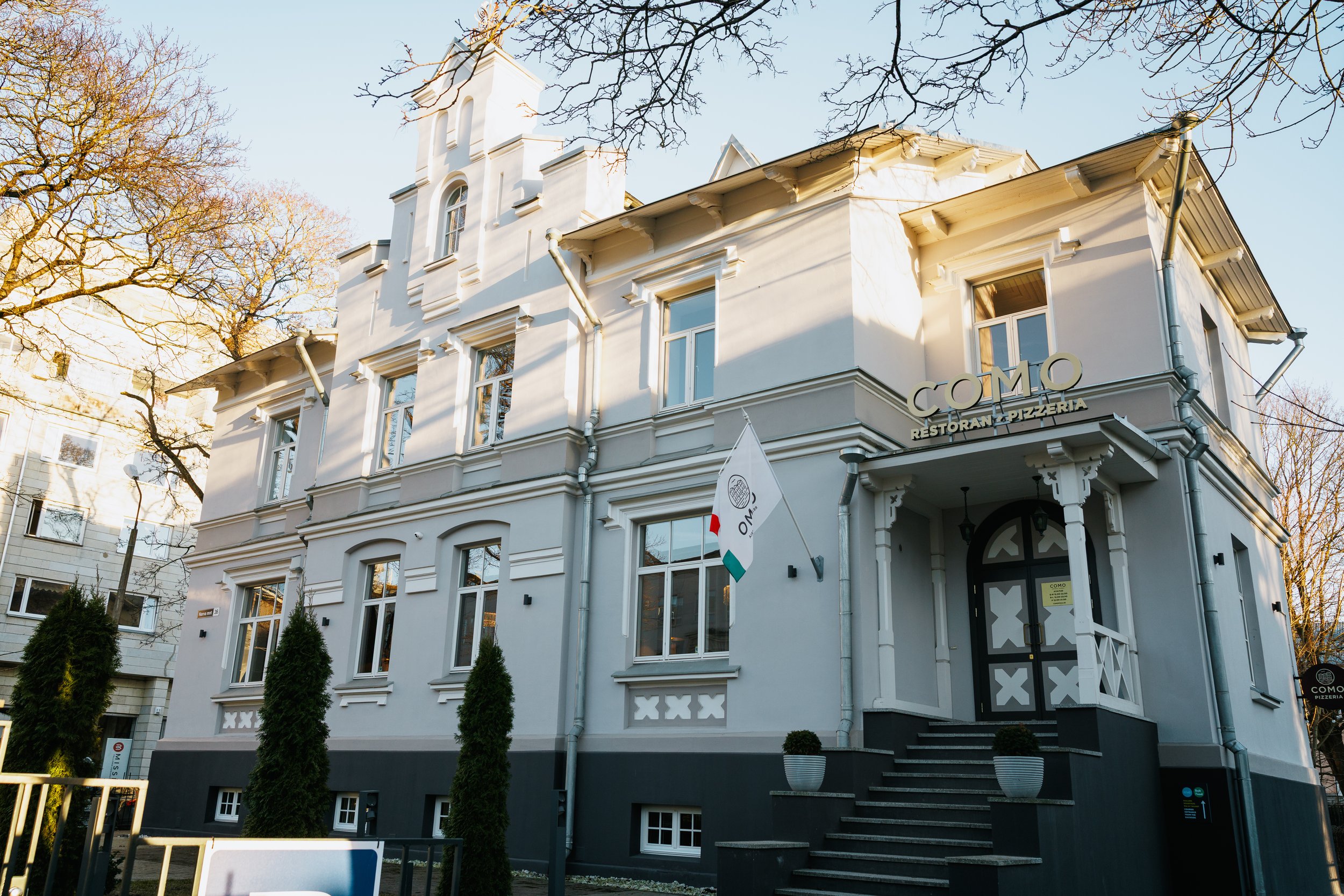 A white multi-story building with decorative architectural details, with a sign that says 'COMO RESTORAN & PIZZERIA' on the front, and a flag with the restaurant's logo hanging near the entrance. There are stairs leading up to the door, and tall ever