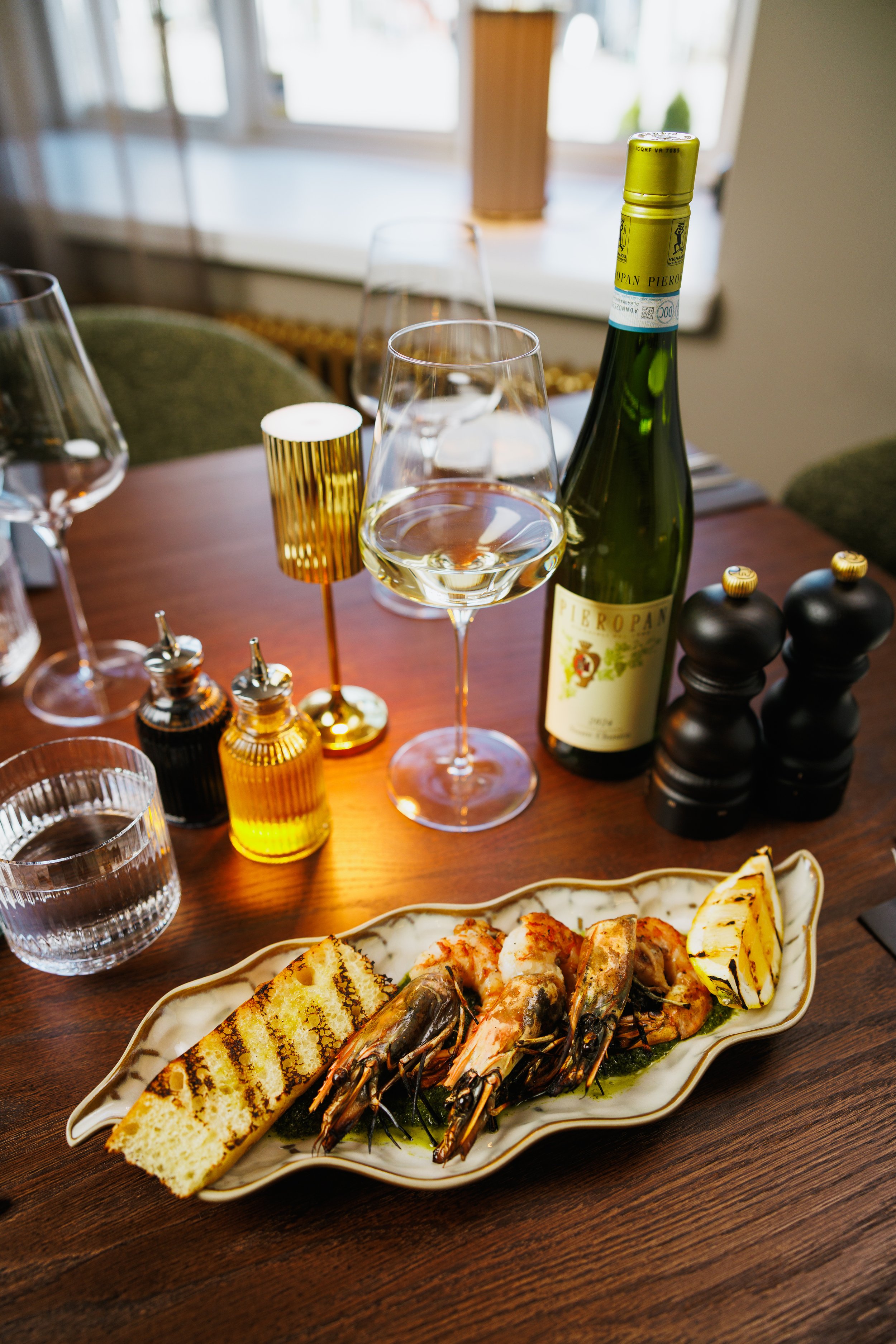 A dinner table set with a plate of grilled shrimp, bread, a glass of white wine, a wine bottle, and various condiments in small bottles, with a window in the background.