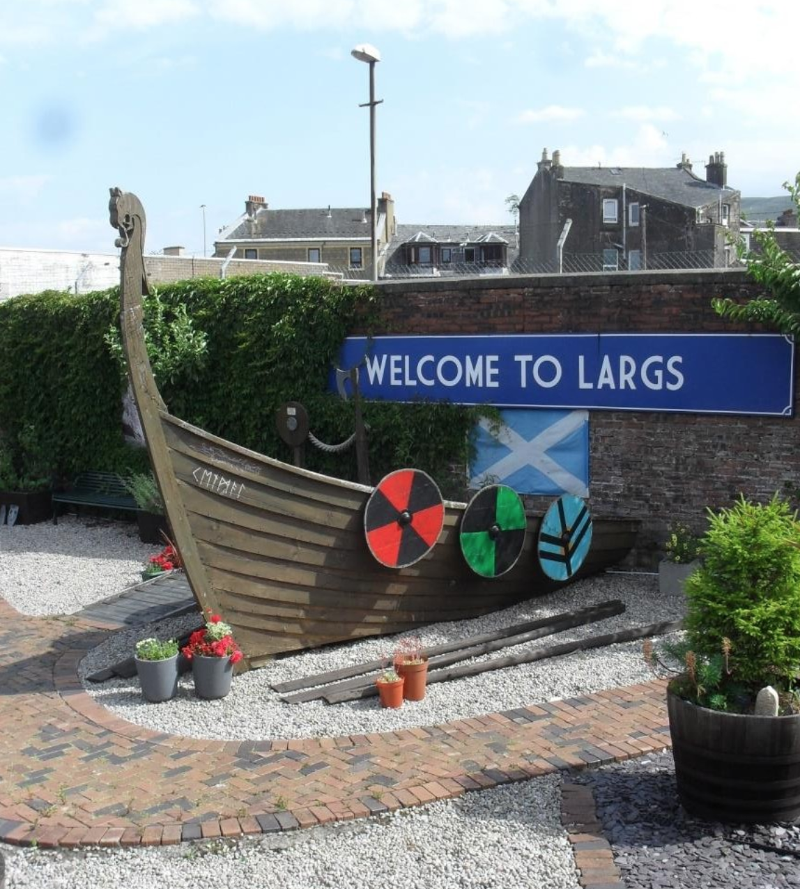 Decorative Viking ship replica with colorful shields and potted plants in front of a sign that says "Welcome to Largs," with buildings and a cloudy sky in the background.