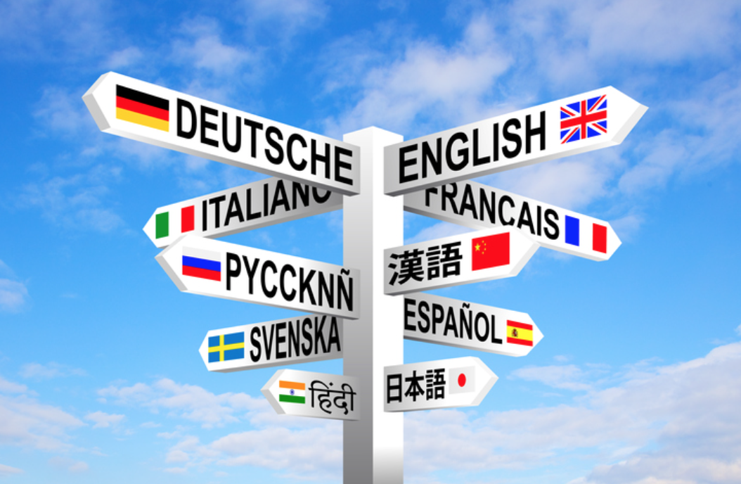 A signpost with arrows pointing in different directions, displaying language names in various scripts and flags against a blue sky background.