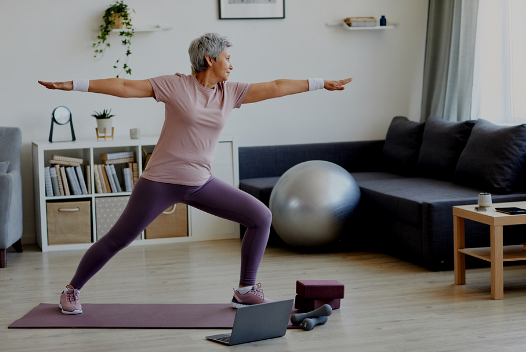 An older woman doing yoga in a living room, standing in warrior pose on a yoga mat with workout equipment nearby.