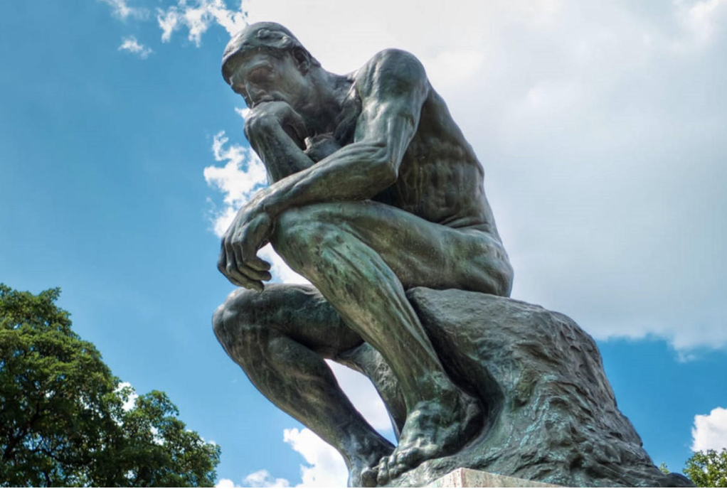 Bronze statue of a man sitting on a rock with his chin resting on his hand, deep in thought, with a blue sky and some trees in the background.