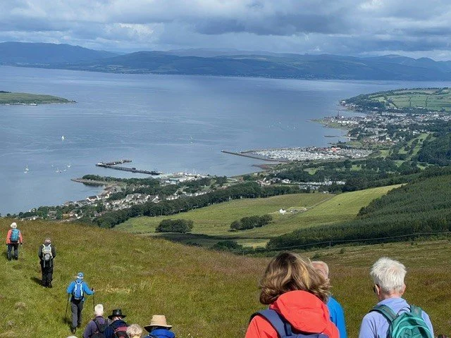 Group of people hiking on a green hillside overlooking a harbor with boats and a town, with mountains in the distance under cloudy skies.