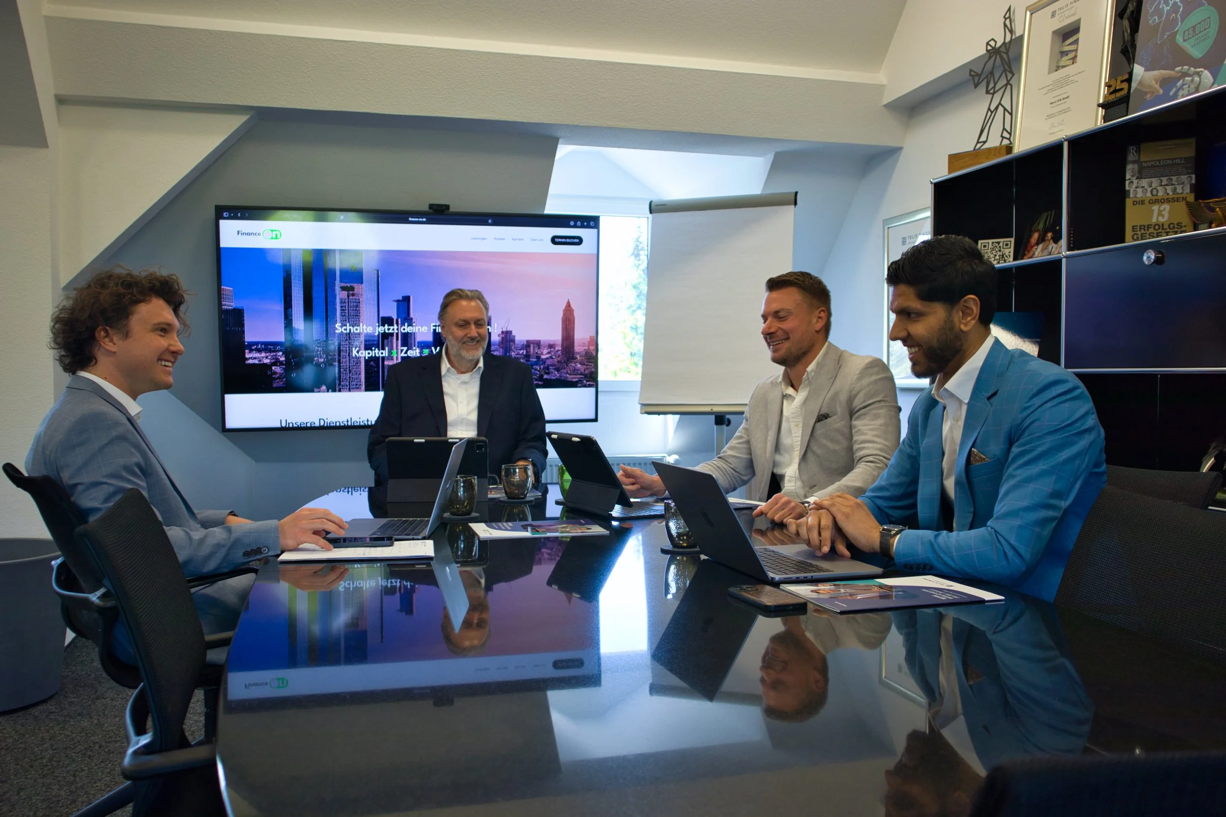 Vier Männer in Business-Formalität sitzen in einem Konferenzraum und unterhalten sich, vor einem großen Bildschirm mit der Finance On Website. Sie haben Laptops, Tablets und Dokumente vor sich.
