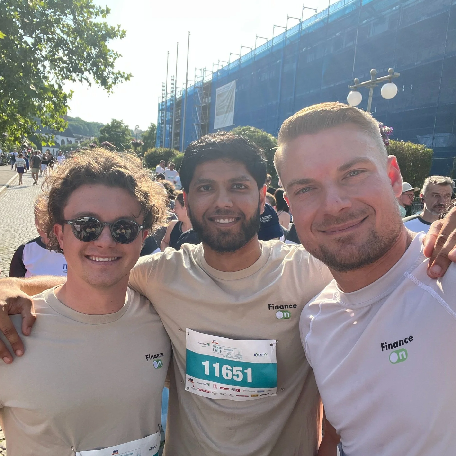 Drei junge Männer beim Firmen-Lauf-Event in Saarbrücken 2025, lächelnd und mit Arme um die Schultern, tragen beige T-Shirts mit dem 'Finance On'-Logo, im Hintergrund viele weitere Läufer.