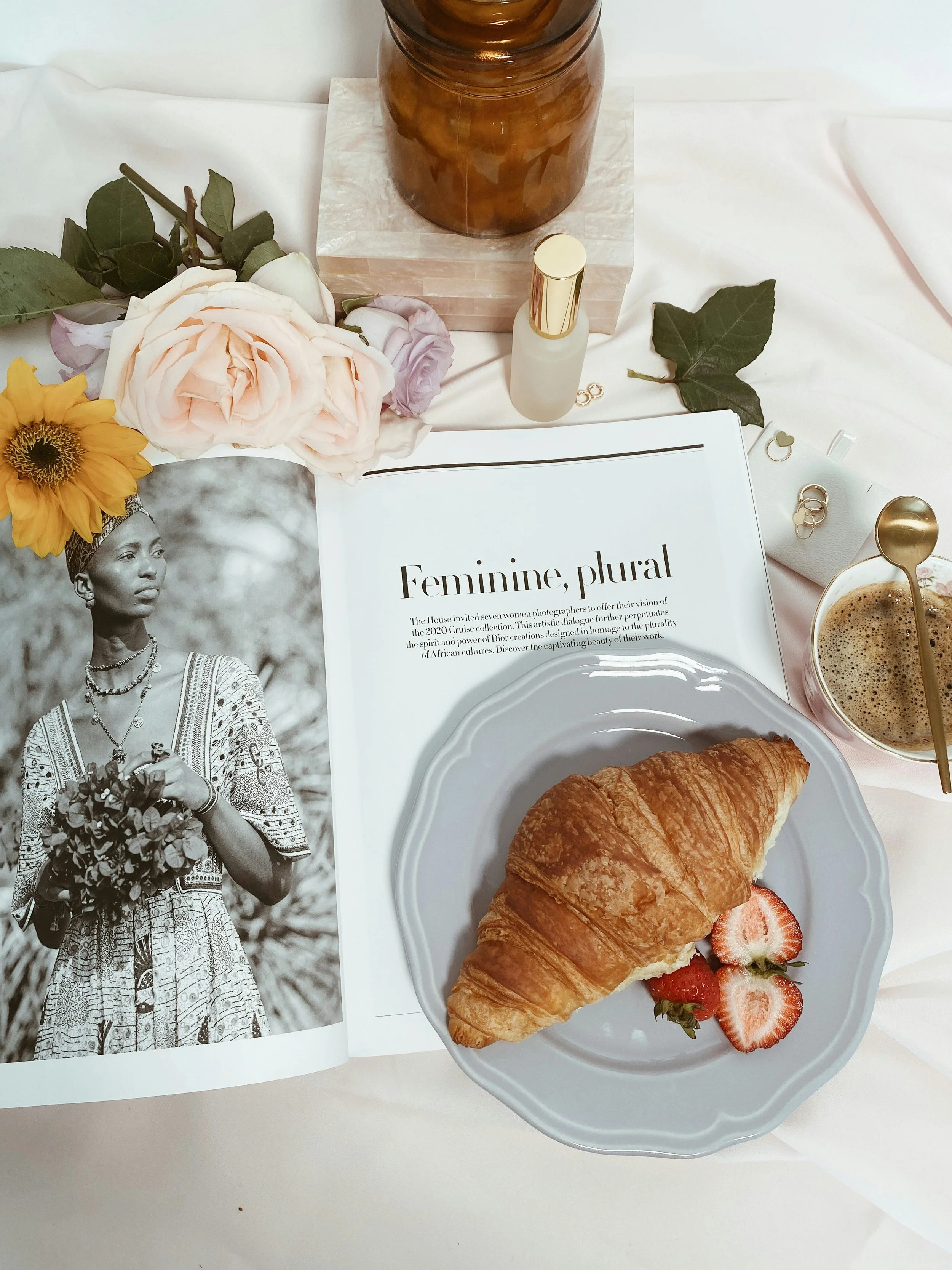 A breakfast scene with a croissant and strawberries on a plate, an open magazine with a black-and-white photo of a woman holding flowers and the title 'Feminine, plural,' a cup of coffee, a jar of honey, flowers, green leaves, and skincare products on a white surface.