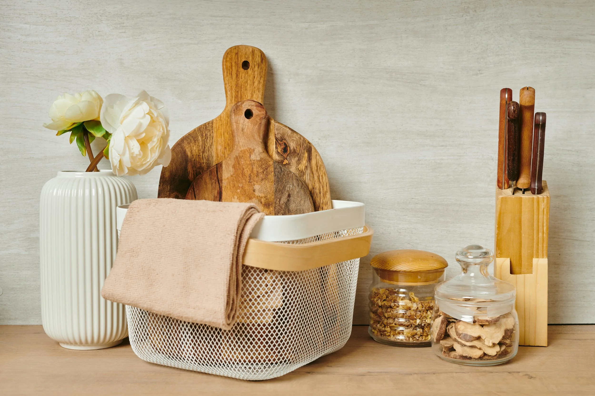 Kitchen countertop with white flowers in a ribbed vase, wooden cutting boards, a beige dish towel, a white basket, jars of spices or dried herbs, and knife blocks with knives.