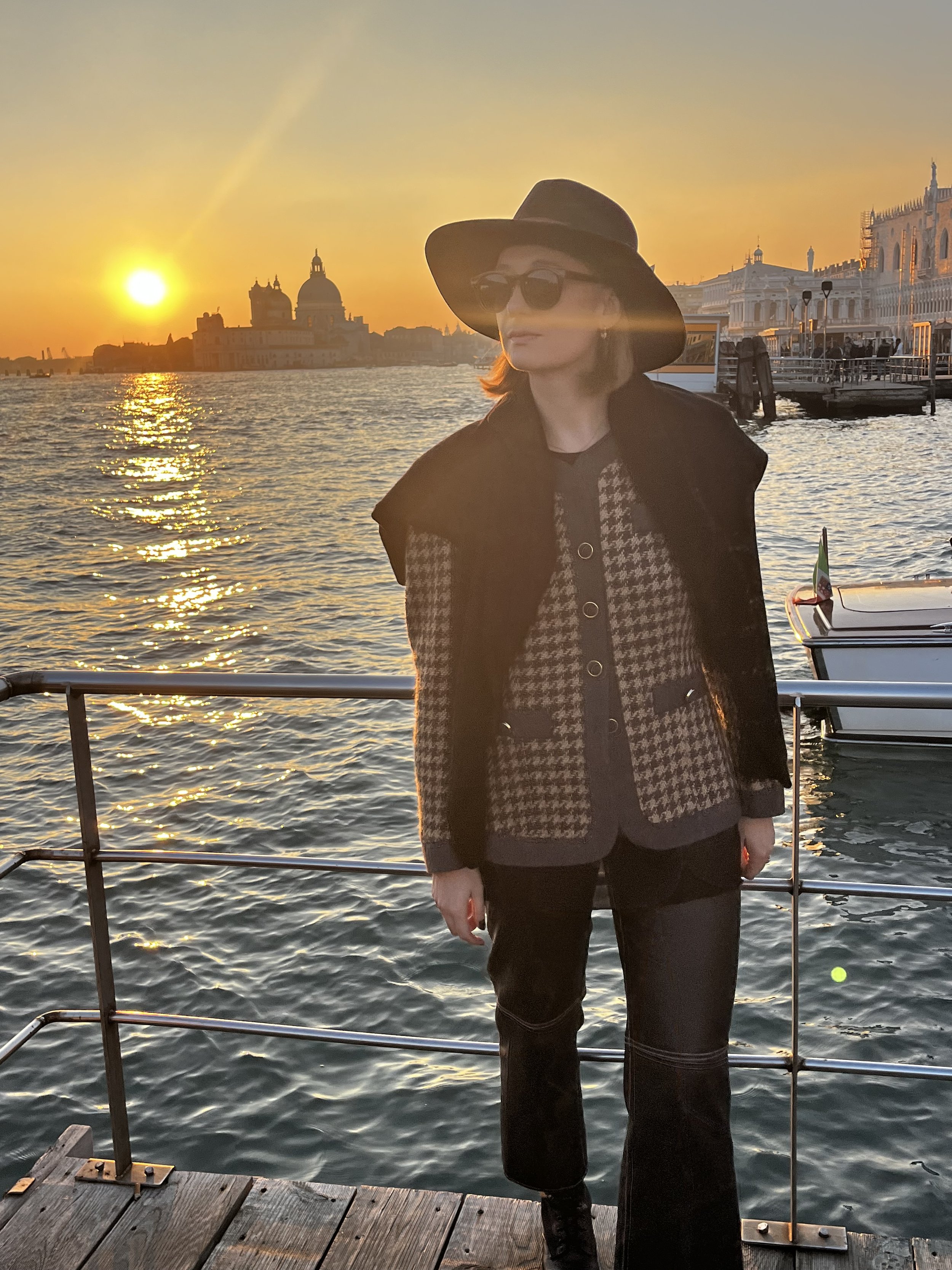 A woman standing on a dock by the water at sunset, wearing a wide-brimmed hat, sunglasses, a houndstooth blazer, and black pants, with a cityscape and a domed building in the background.