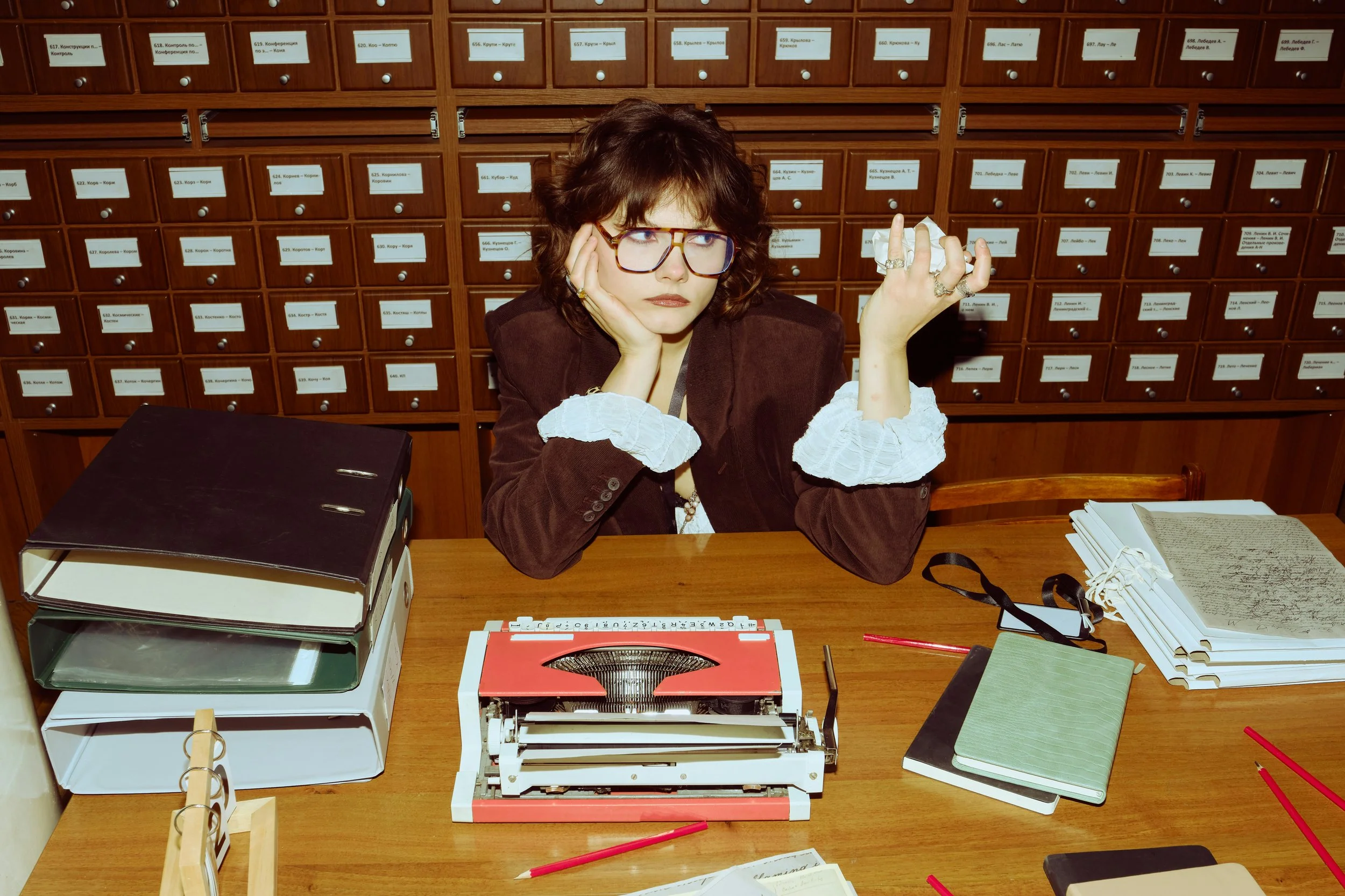 A woman sitting at a desk with a frustrated and bored expression, surrounded by binders, notebooks, typewriter, and scattered papers, in front of a wall of small labeled drawers.