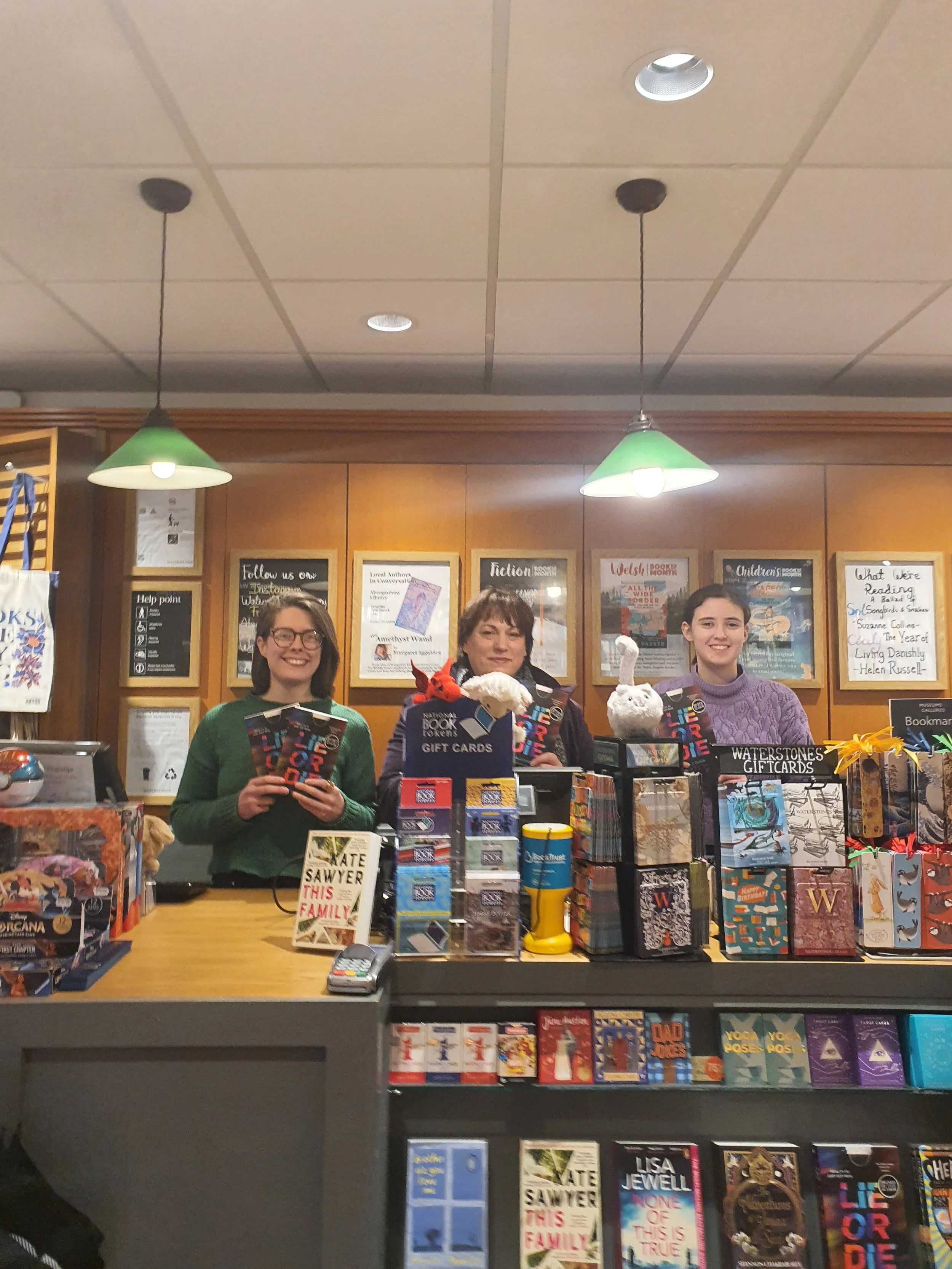 Three women standing behind a bookstore checkout counter, smiling at the camera. The counter is filled with books, gift cards, and plush toys, with wooden wall decor and framed posters in the background.