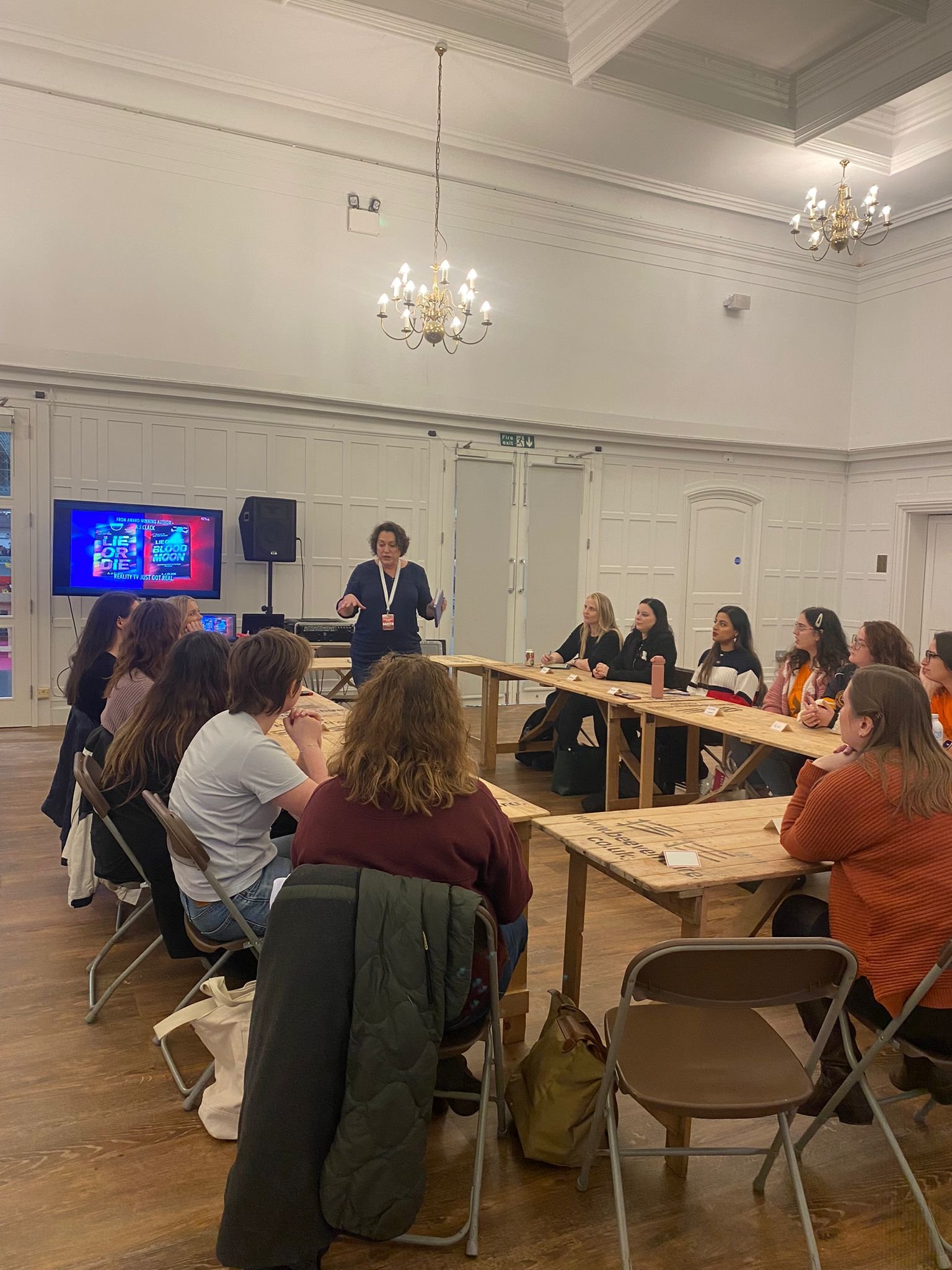A woman standing and speaking to a group of women seated at wooden tables in a room with white paneled walls and chandeliers, with a screen displaying a presentation titled 'Lie for You'.