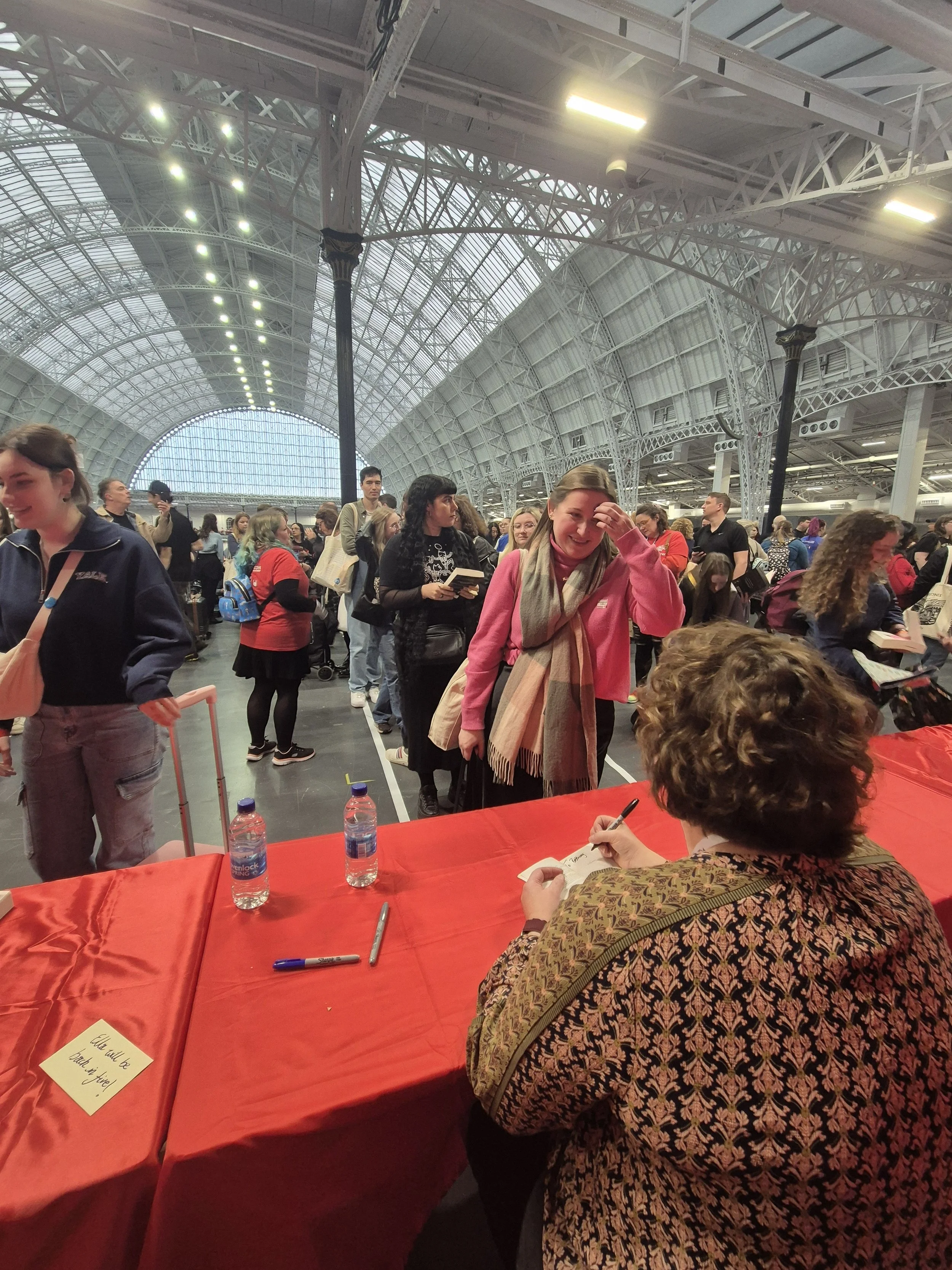 A woman with curly hair and a patterned shirt is sitting at a table signing a book or paper. There are water bottles and pens on the table. A line of people is waiting, and the woman in the pink jacket is smiling and touching her forehead while standing in line inside a large, arched, glass-and-steel building.