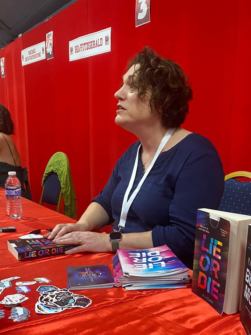 A woman with curly hair and a dark blue top sitting at a table with books and stickers, at a red booth labeled 'BEA FITZGERALD' with some signs and a water bottle nearby.