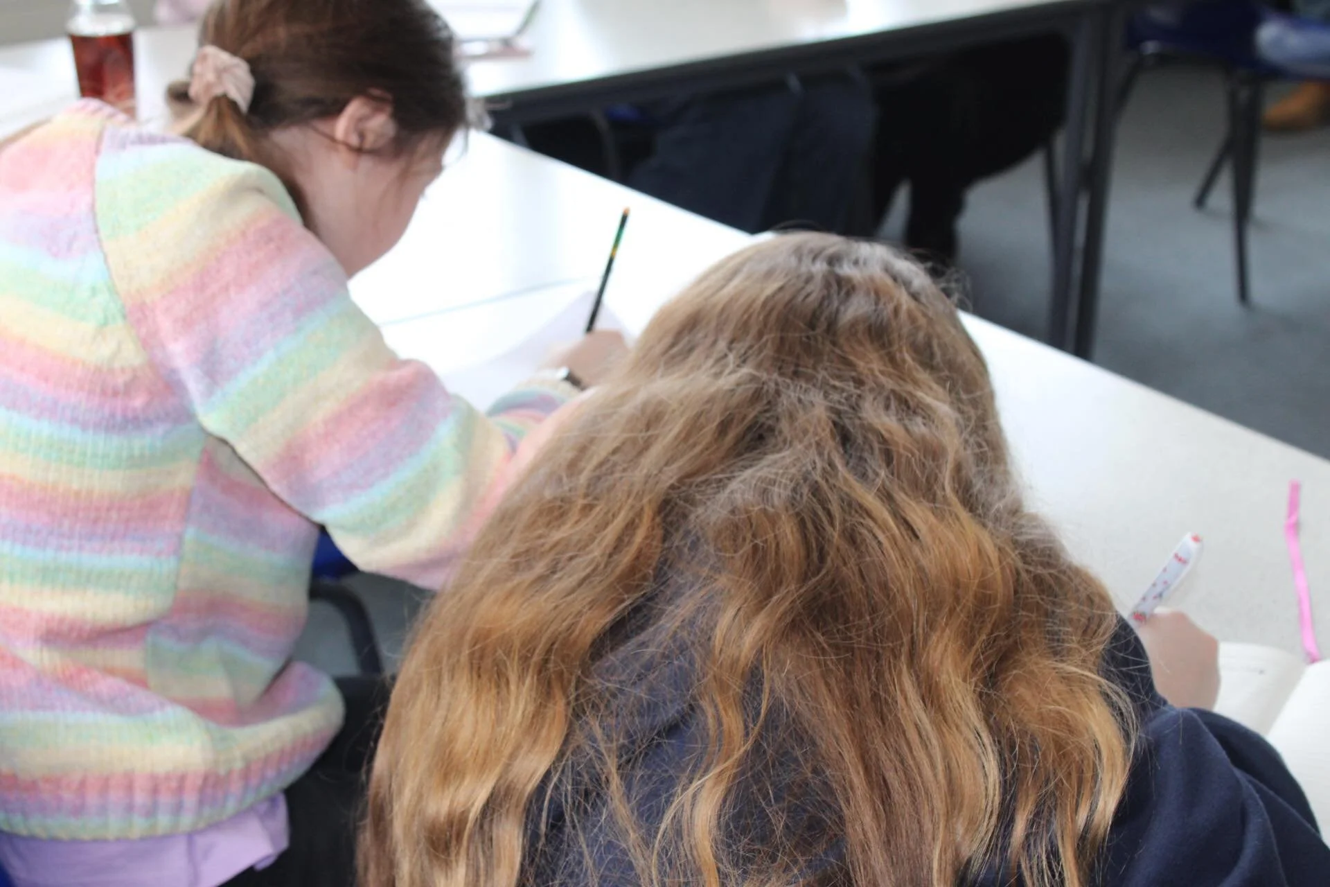 Two young women are sitting at a desk, focused on writing or drawing, with one wearing a pastel rainbow-striped sweater and the other with long red hair.
