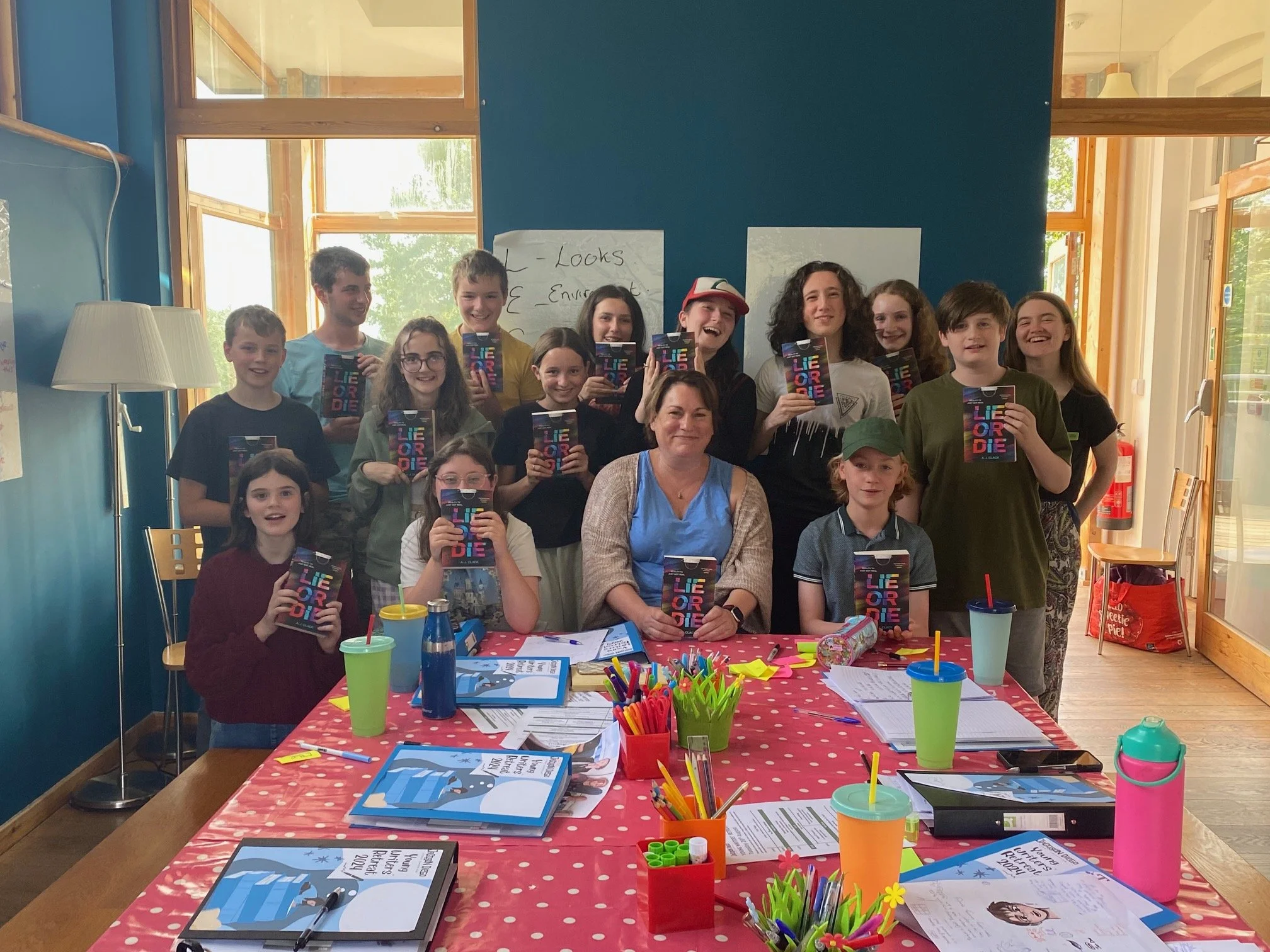 A group of children and a teacher gathered around a table in a classroom, holding books titled 'Liar, Liar.' The table has school supplies and colorful cups, with large windows in the background letting in natural light.