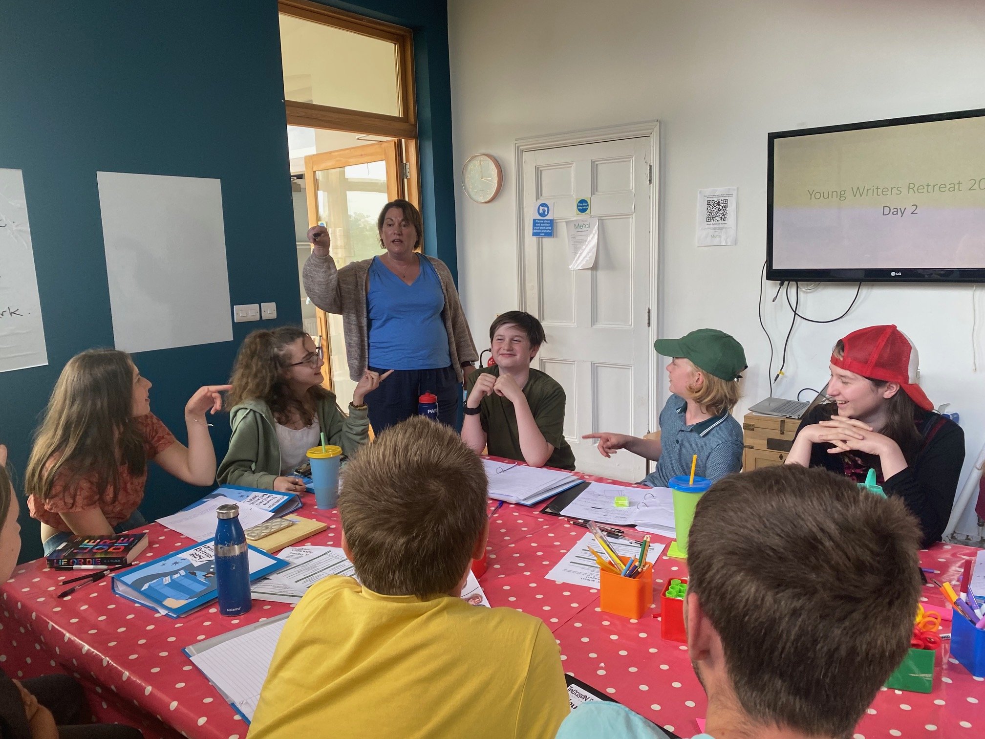 A group of young people and a woman are sitting around a table during a workshop or class. The woman, standing, is speaking while the others listen and interact. The table has notebooks, papers, and colorful stationery. There is a large screen on the