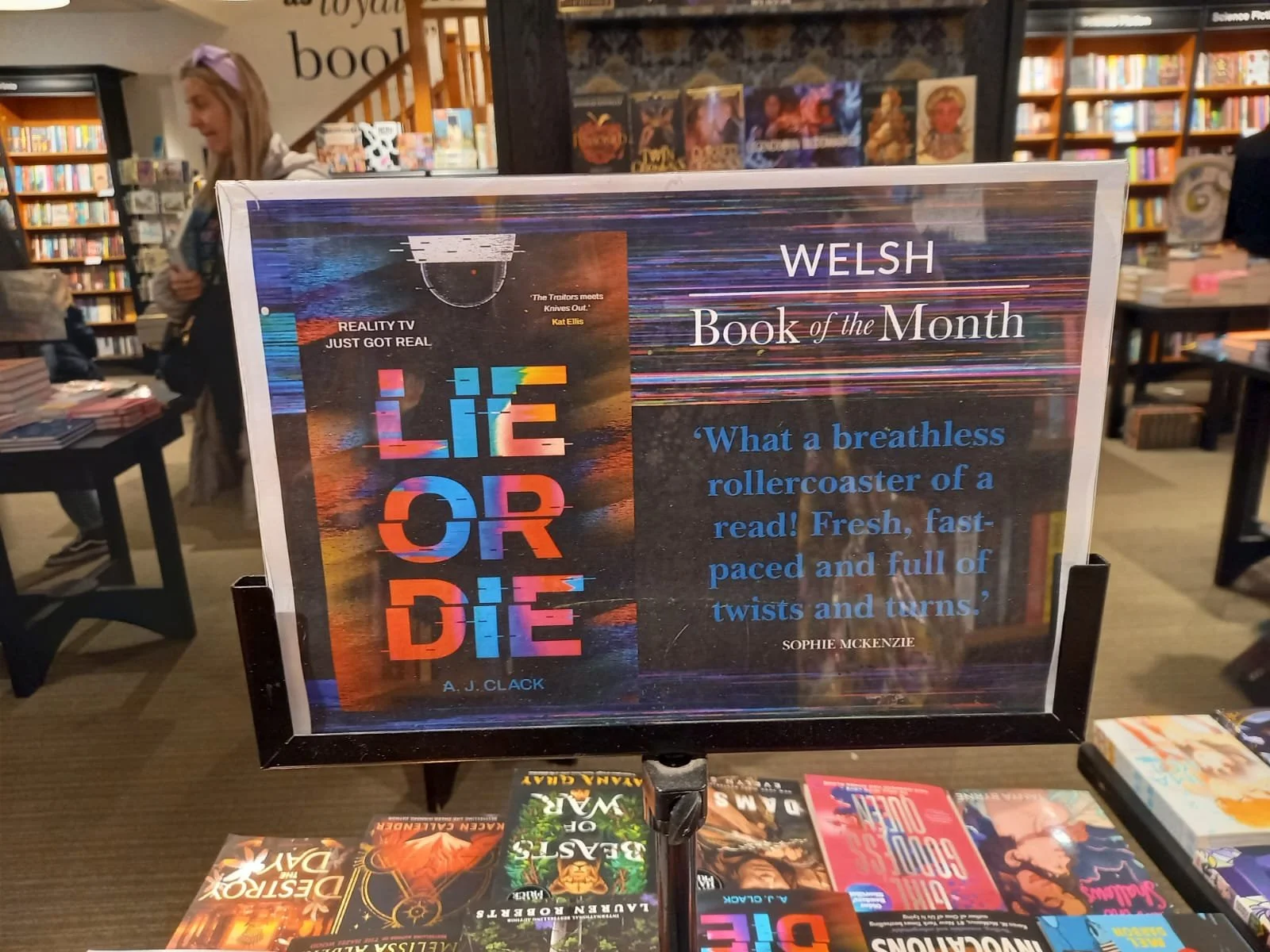 A bookstore display sign for the Welsh Book of the Month, featuring the book "Life or Die" by A. J. Clack, with a quote from Sophie McKenzie praising the book as a breathless rollercoaster of a read.