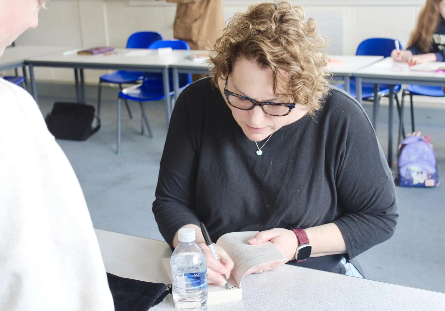 A woman with curly hair and glasses reading a book at a table with a water bottle, in a classroom with blue chairs and other students in the background.