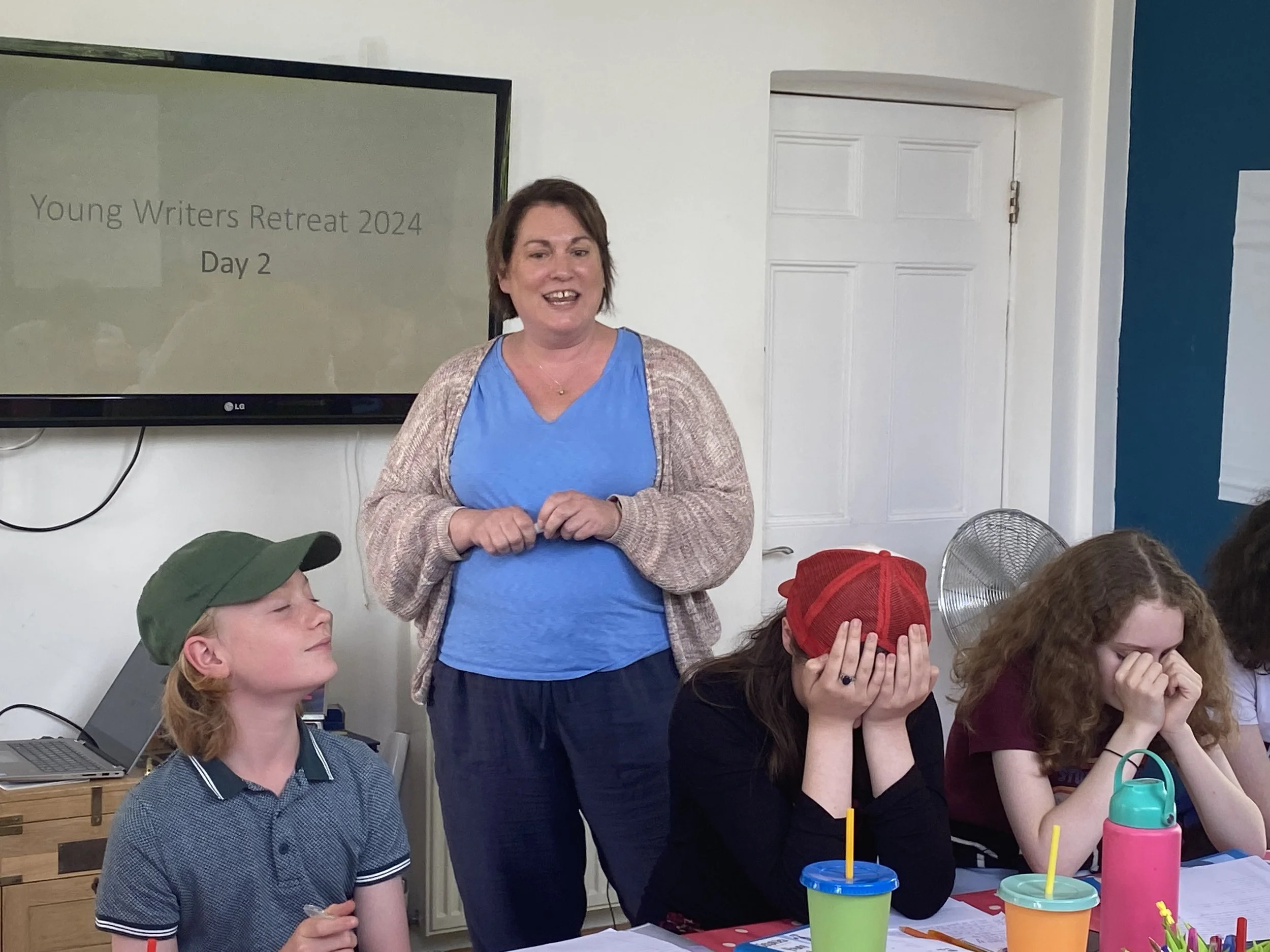 A woman standing and talking in front of a group of young people at a retreat, with a screen behind her displaying 'Young Writers Retreat 2024, Day 2'.