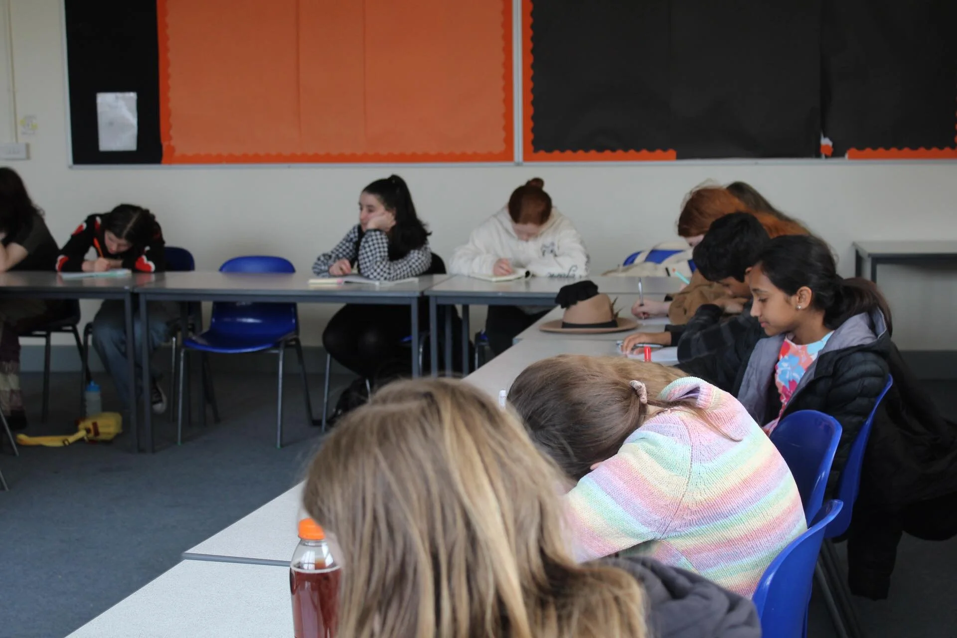 Students sitting at desks in a classroom, engaged in writing or resting, with some wearing jackets and hats.