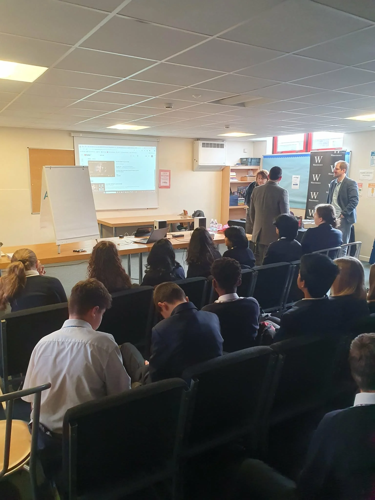 People attending a presentation or lecture in a classroom or conference room with a projected screen, whiteboard, and banners.
