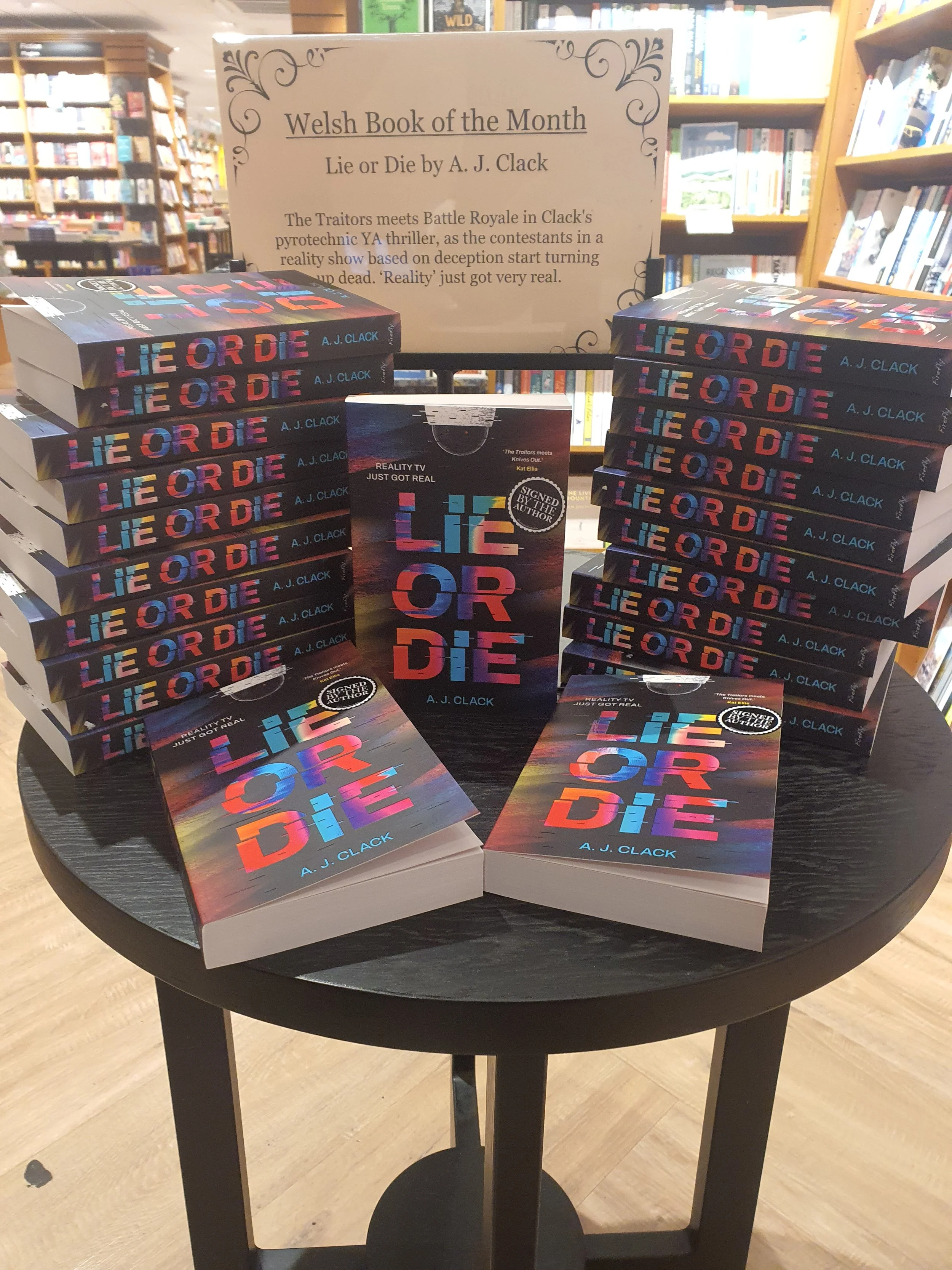 Display of multiple copies of the book 'Lie or Die' by A. J. Clack on a black round table in a bookstore, with a sign behind them promoting the book as the Welsh Book of the Month.
