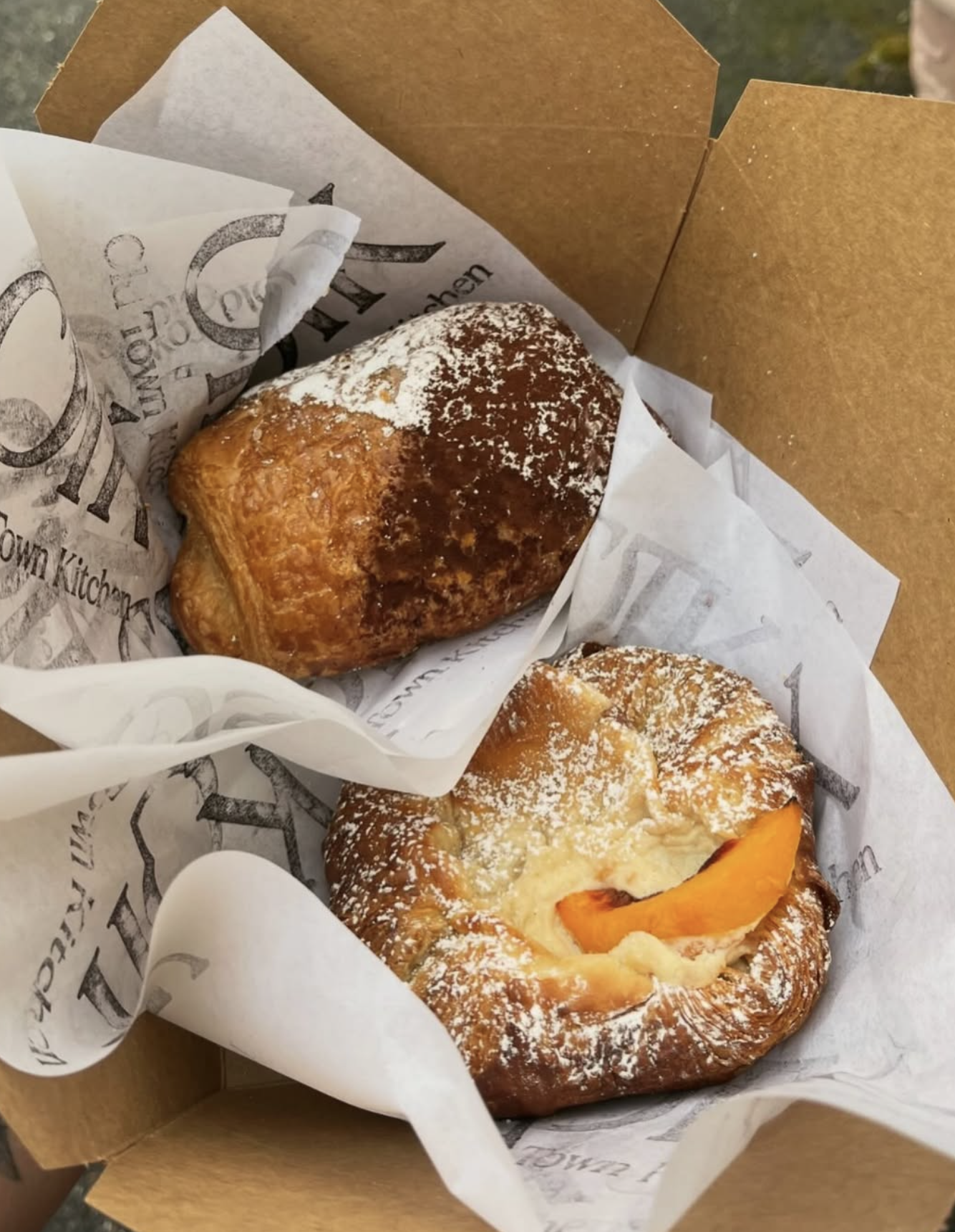 Two pastries in a brown cardboard box with decorative paper lining. The pastry on the left appears to be an apple turnover dusted with powdered sugar. The pastry on the right looks like a Danish with apricot filling, also dusted with powdered sugar.