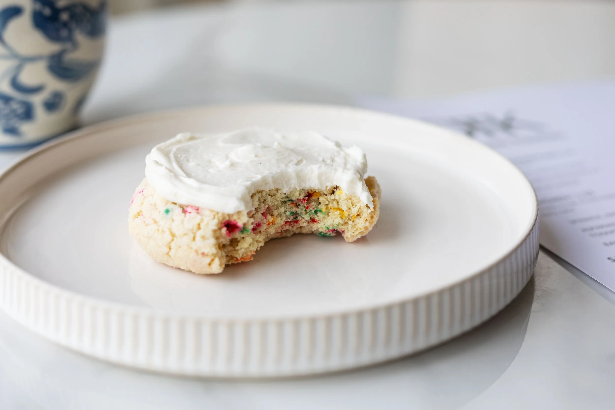 A partially eaten sugar cookie with colorful sprinkles and white frosting on top, resting on a white plate.