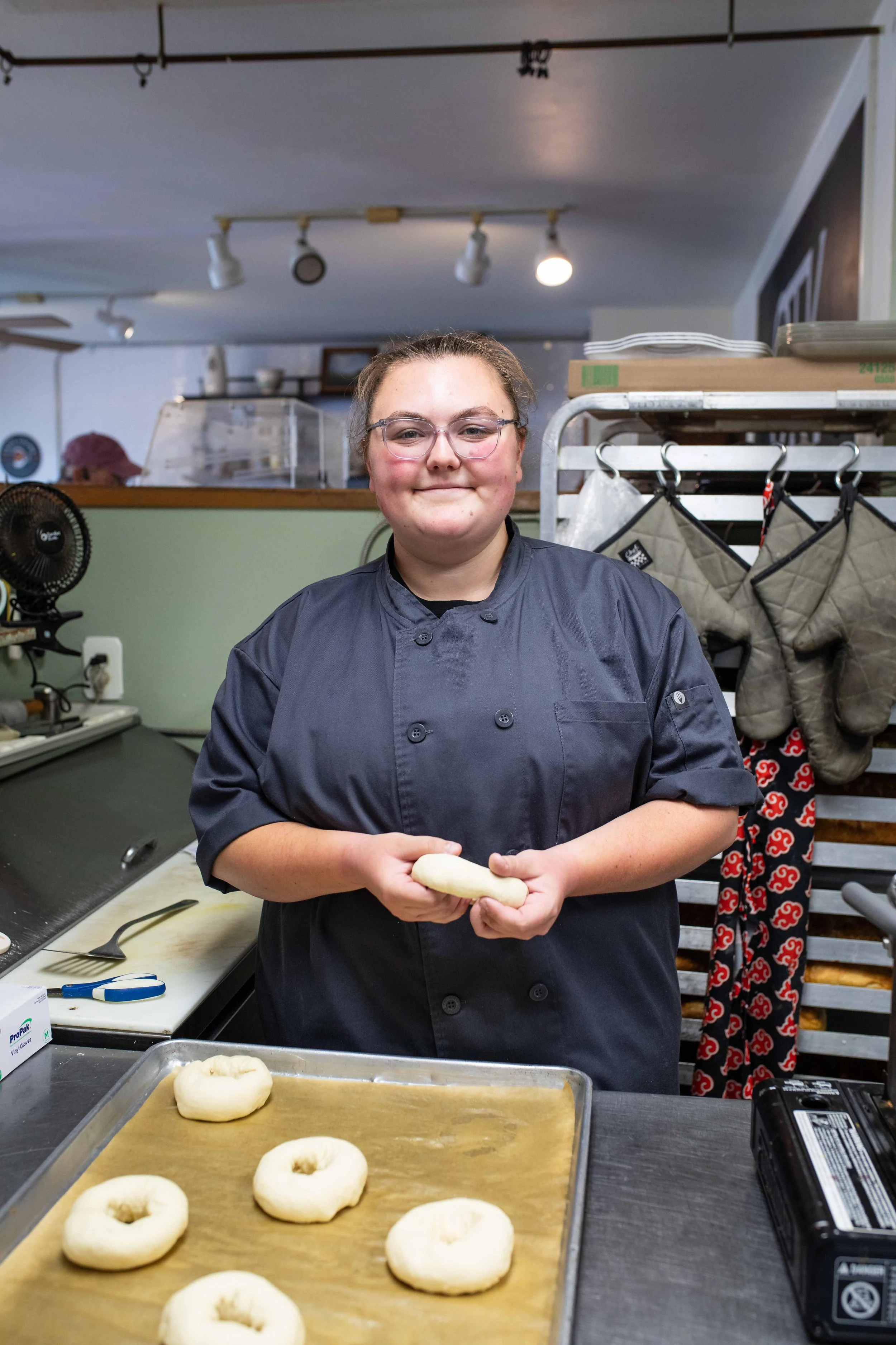 A woman wearing glasses and a black chef's coat stands in a kitchen, holding a piece of dough. There is a tray with four unbaked bagels on a baking sheet in the foreground. The background shows kitchen equipment, a rack with hanging oven mitts, and a person in a pink hat working in the kitchen.